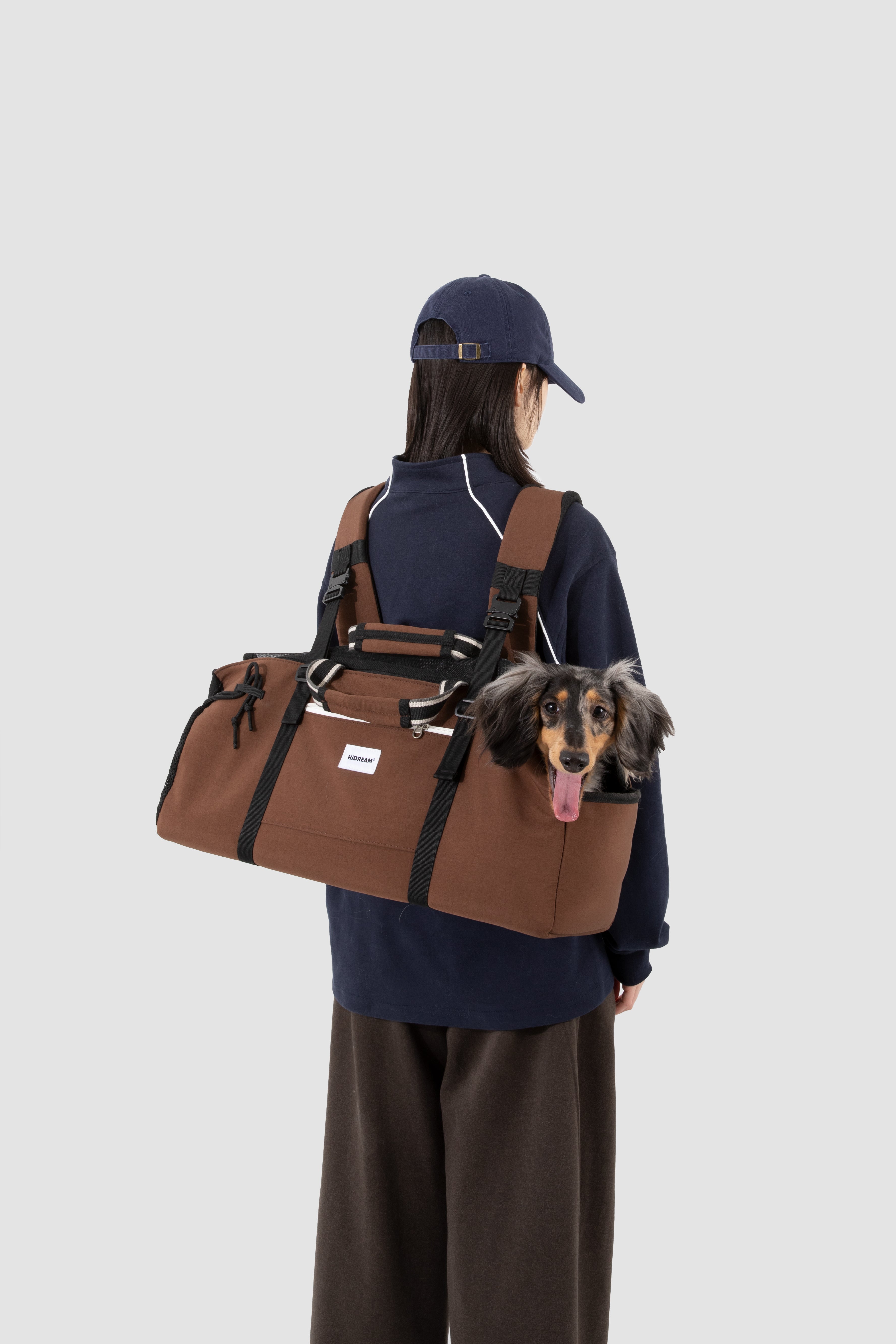 A person wearing a navy blue cap, navy sweatshirt, and brown pants is shown from the back, carrying a brown Dachshund carrier duffel bag on their shoulder. A Dachshund is peeking out of the top of the bag.