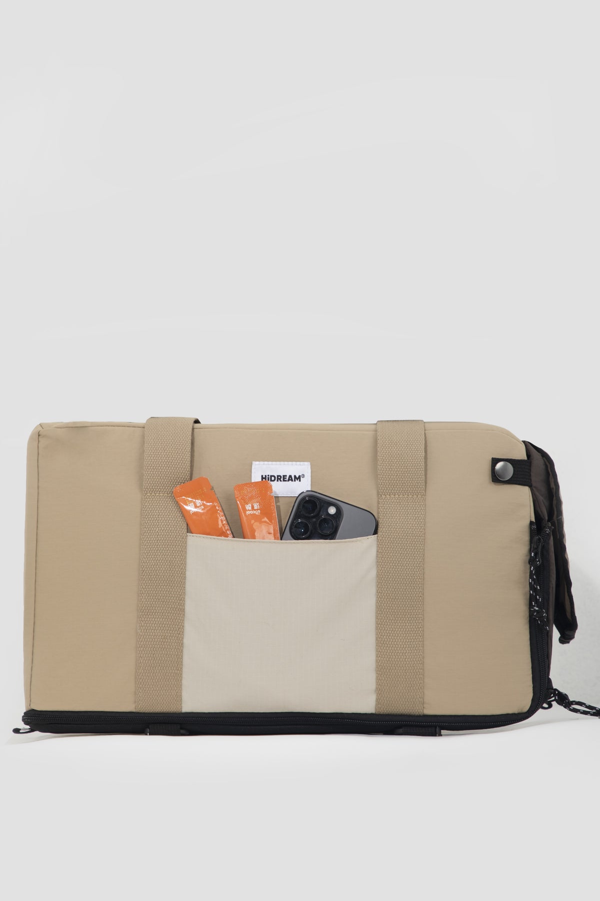 A beige canvas travel pouch or small duffel bag with black trim, shown against a light gray background. The front pocket is open, displaying orange and white items inside.