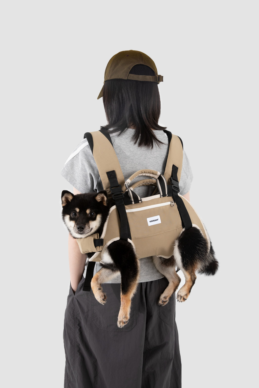 A person shown from behind wearing a tan dog carrier backpack with a small black and tan dog inside, against a plain light gray studio background.