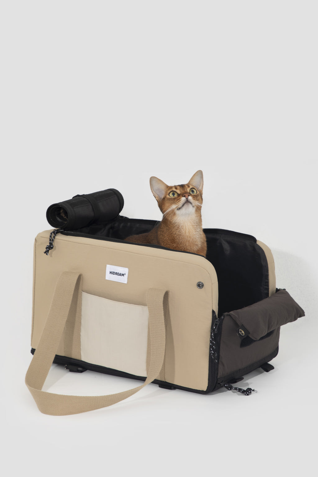 A tan and black pet carrier bag with a small cat sitting inside, photographed on a clean light gray studio background.