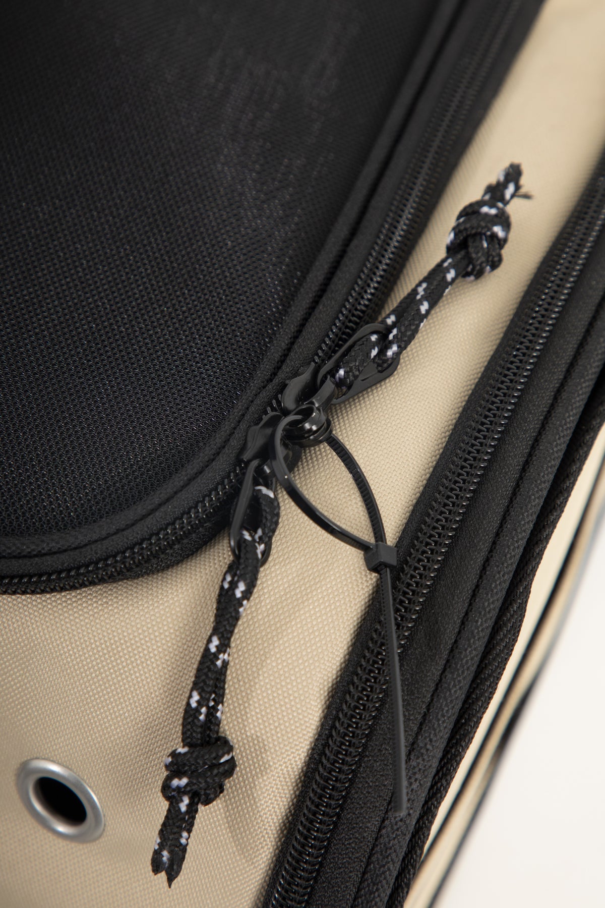 Close-up of a beige and black bag with a black zipper, black fabric panel, and a black cord pull tied in a knot, showing a metal grommet on the beige side.