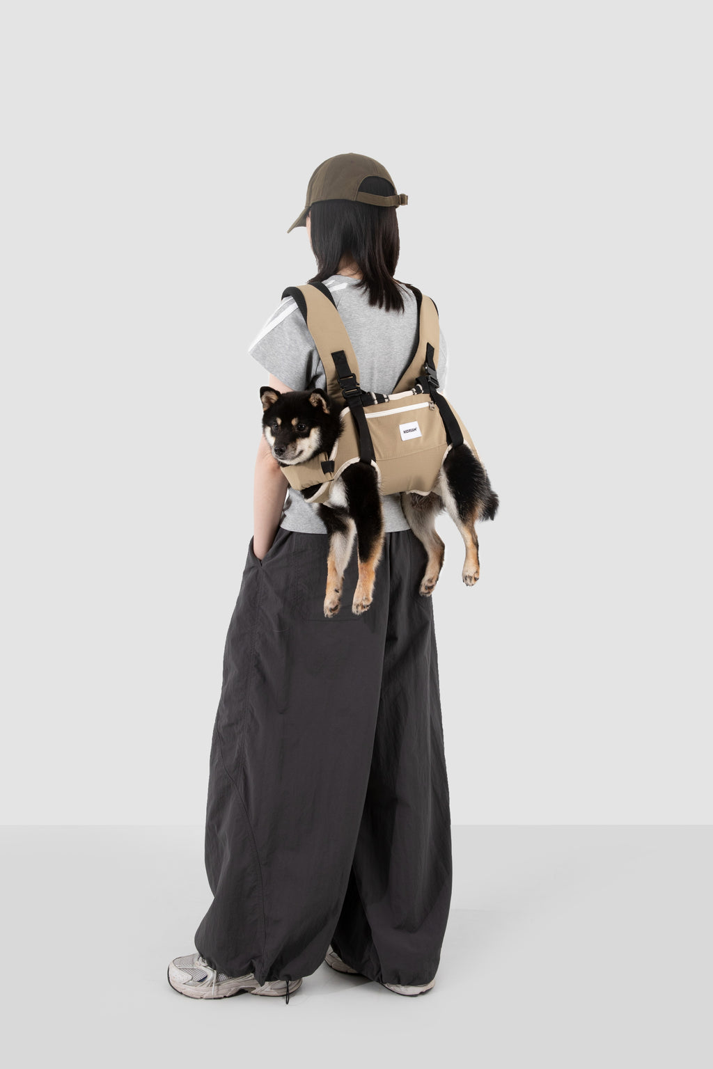 A person wearing a dog carrier backpack with a small black and white dog inside, shown from behind against a light gray studio background.
