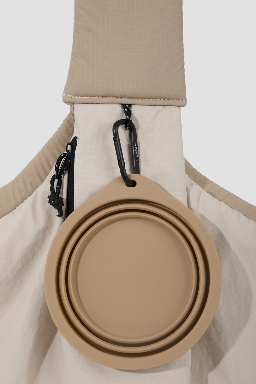 A beige collapsible silicone water bottle holder or cup accessory clipped to the shoulder strap of a light beige bag, shown against a plain light gray background.