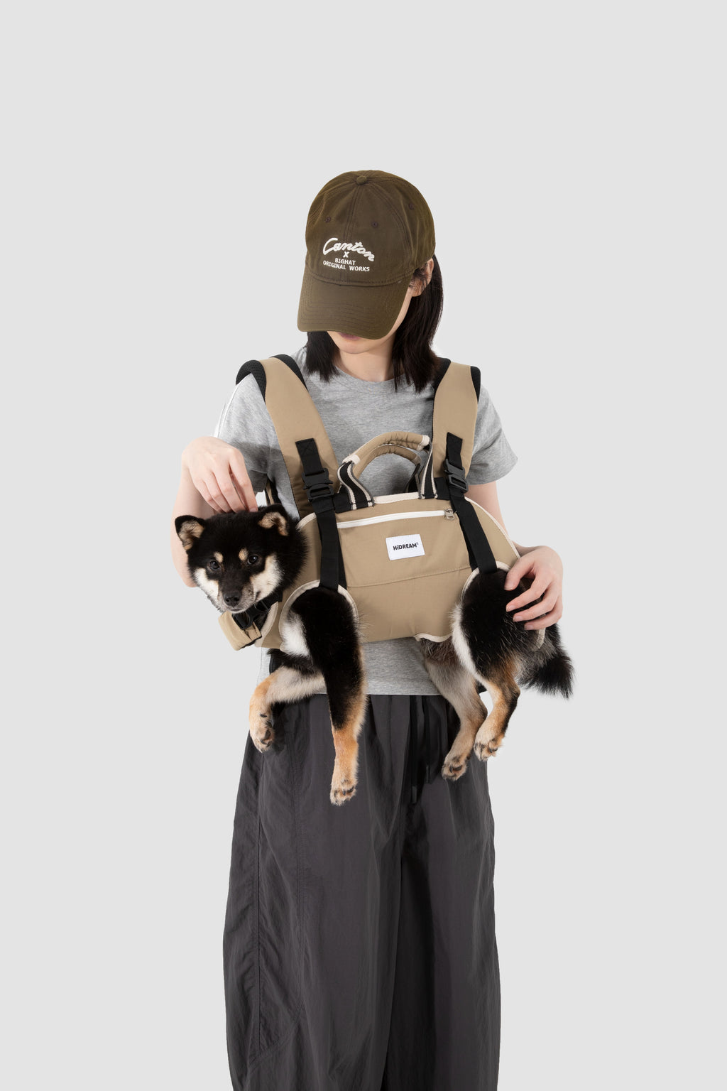 A person wearing a brown cap, gray t-shirt, and dark gray pants models a beige dog carrier backpack with a small black and tan dog sitting inside, against a solid light gray background.