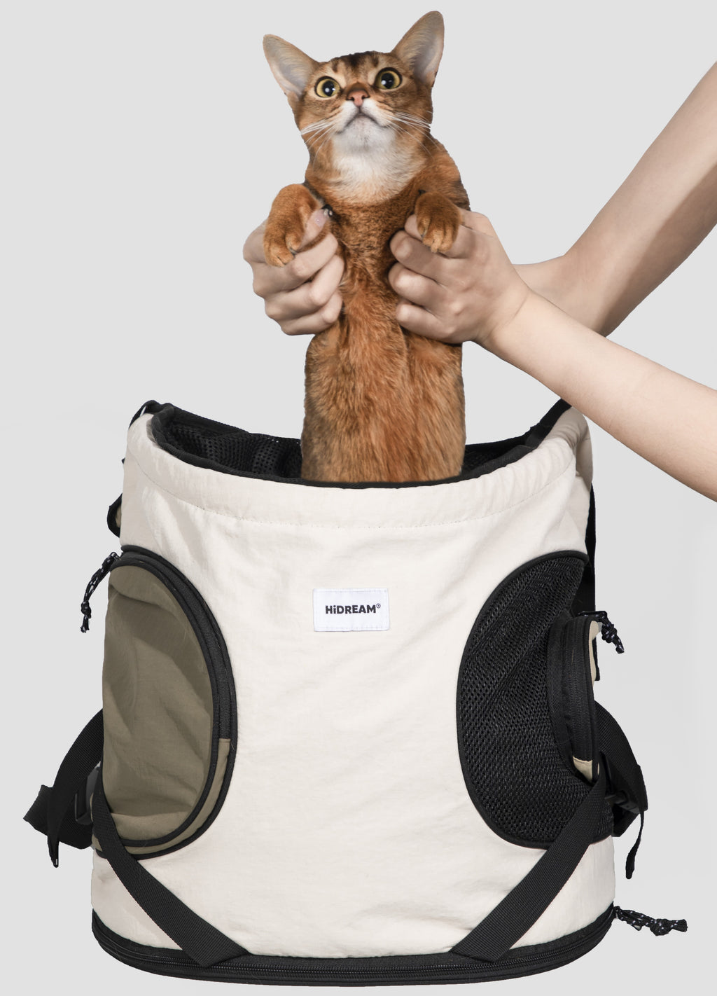A cat backpack carrier with a brown cat inside, held up by a person's hands against a light gray background. The backpack is cream-colored with black mesh and trim, and has a visible 'HIDEAM' brand label.