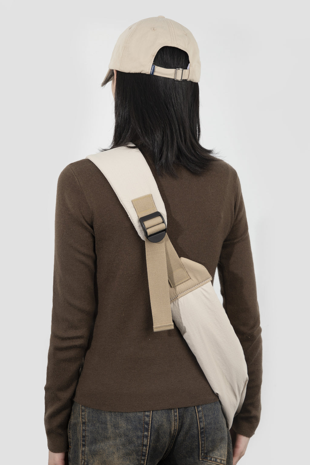 A person shown from the back wearing a brown long-sleeve top, dark jeans, a beige baseball cap, and carrying a beige crossbody bag with a wide strap and black buckle over their shoulder, against a light gray studio background.