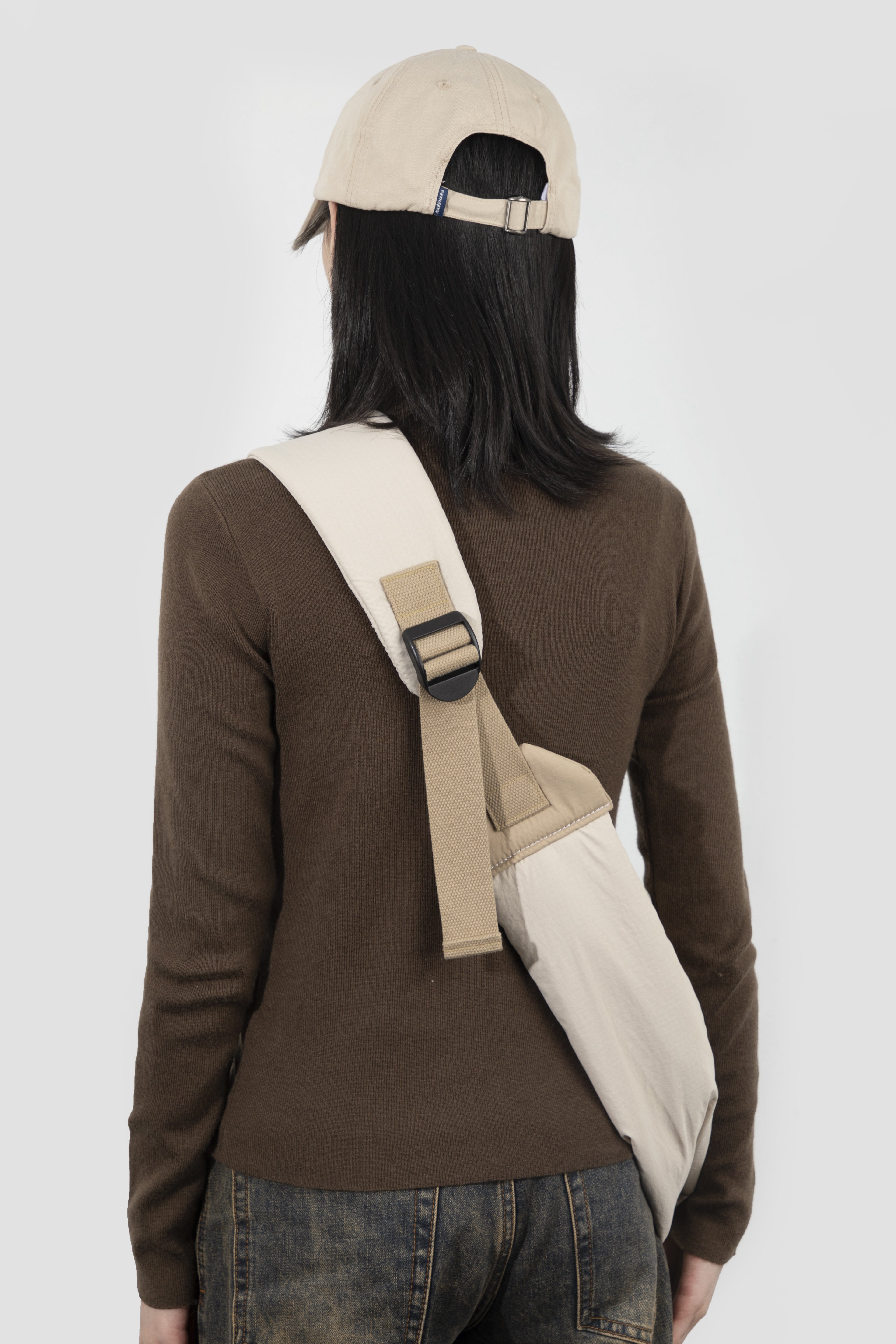 A person shown from the back wearing a brown long-sleeve top, dark jeans, a beige baseball cap, and carrying a beige crossbody bag with a wide strap and black buckle over their shoulder, against a light gray studio background.