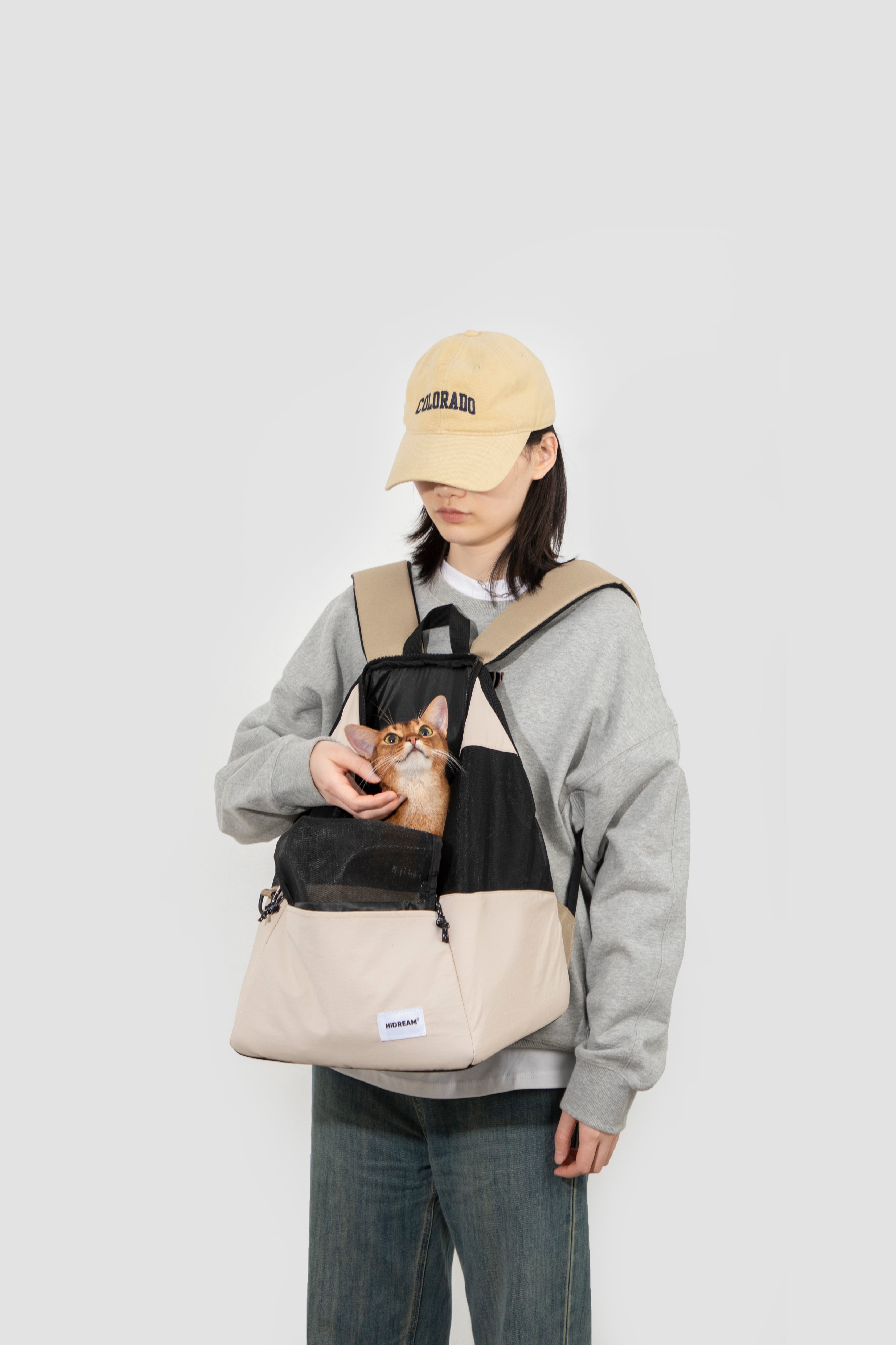 Person holding a cat with a beige backpack on a plain background