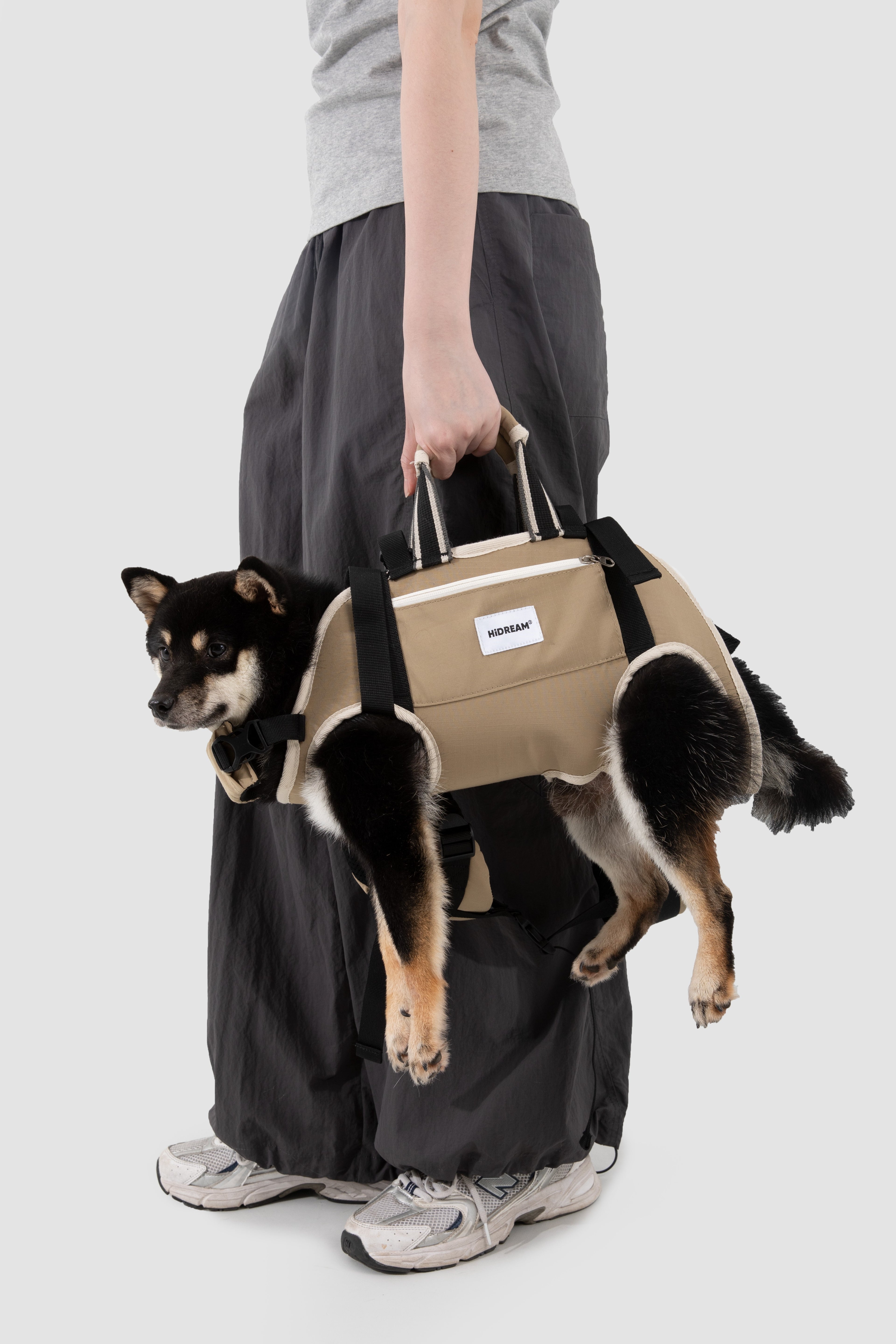 A person wearing loose black pants and a gray top carries a black and tan dog in a beige dog carrier bag with black straps, against a light gray studio background.