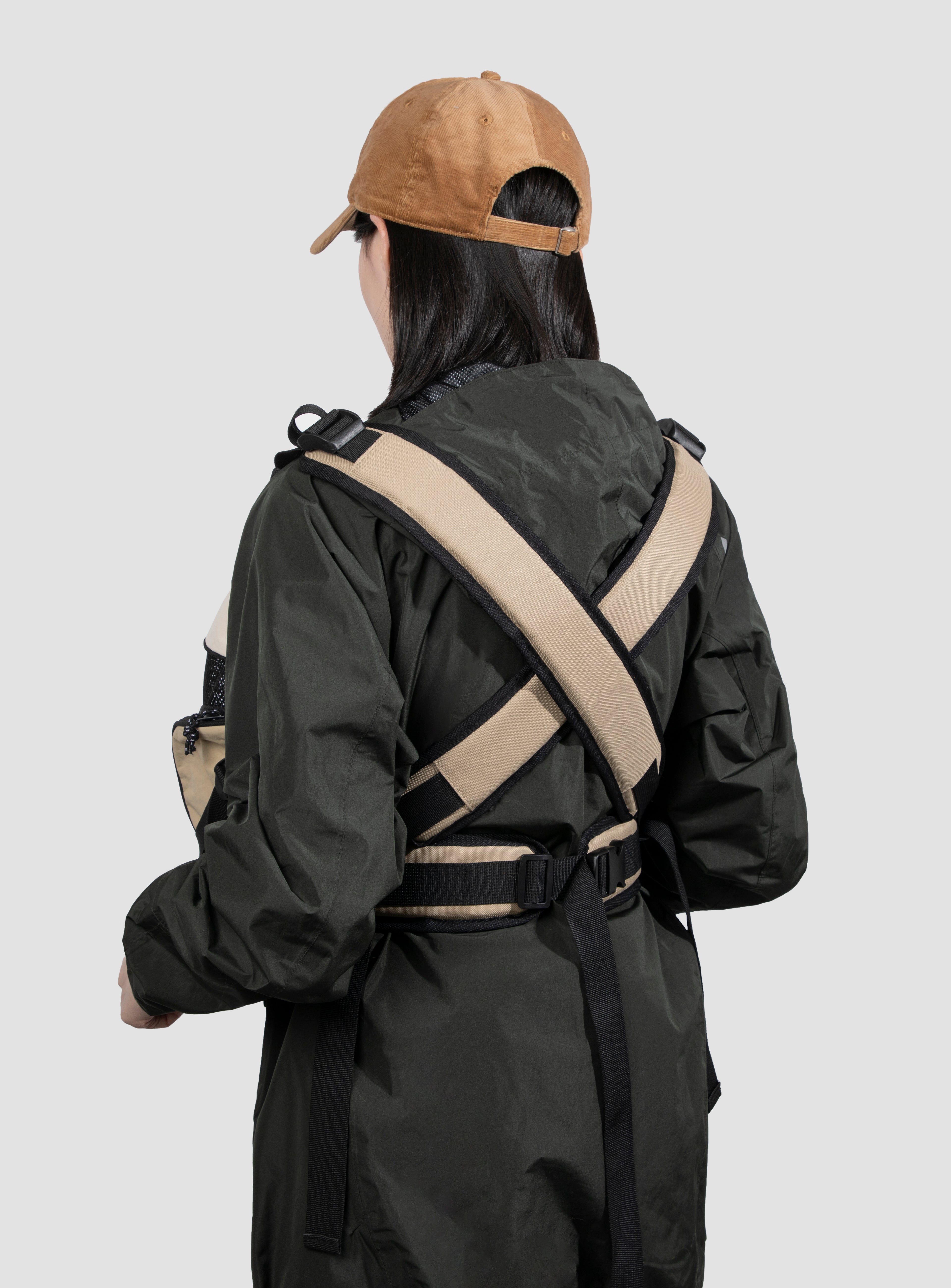 A person shown from the back wearing a black hooded jacket with wide beige crossbody straps and a tan cap, against a plain light gray studio background.