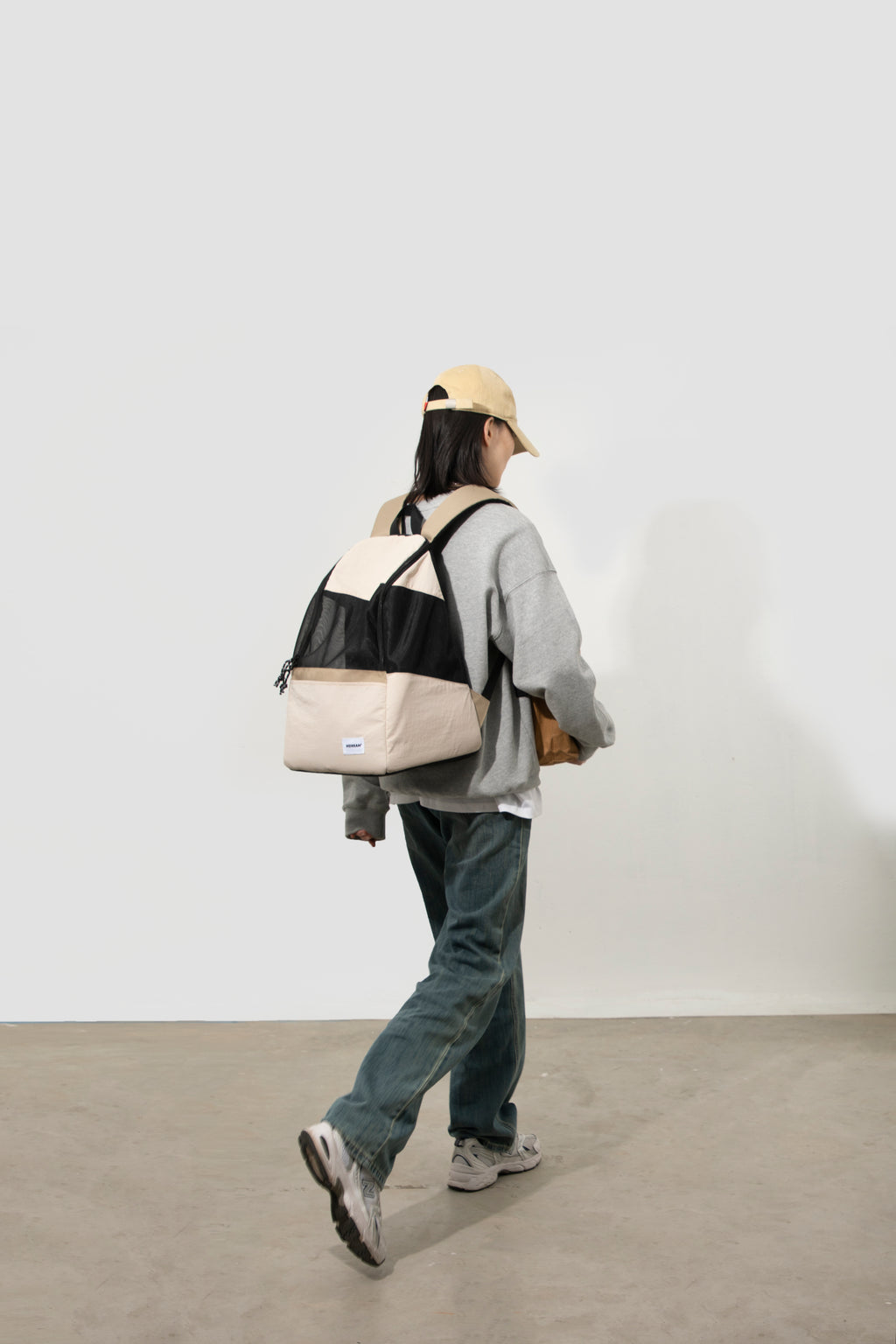 A person walking away in a minimalist studio setting, wearing a light beige baseball cap, a gray oversized sweatshirt, dark gray jeans, and sneakers, while carrying a color-blocked backpack with beige, black, and gray panels.