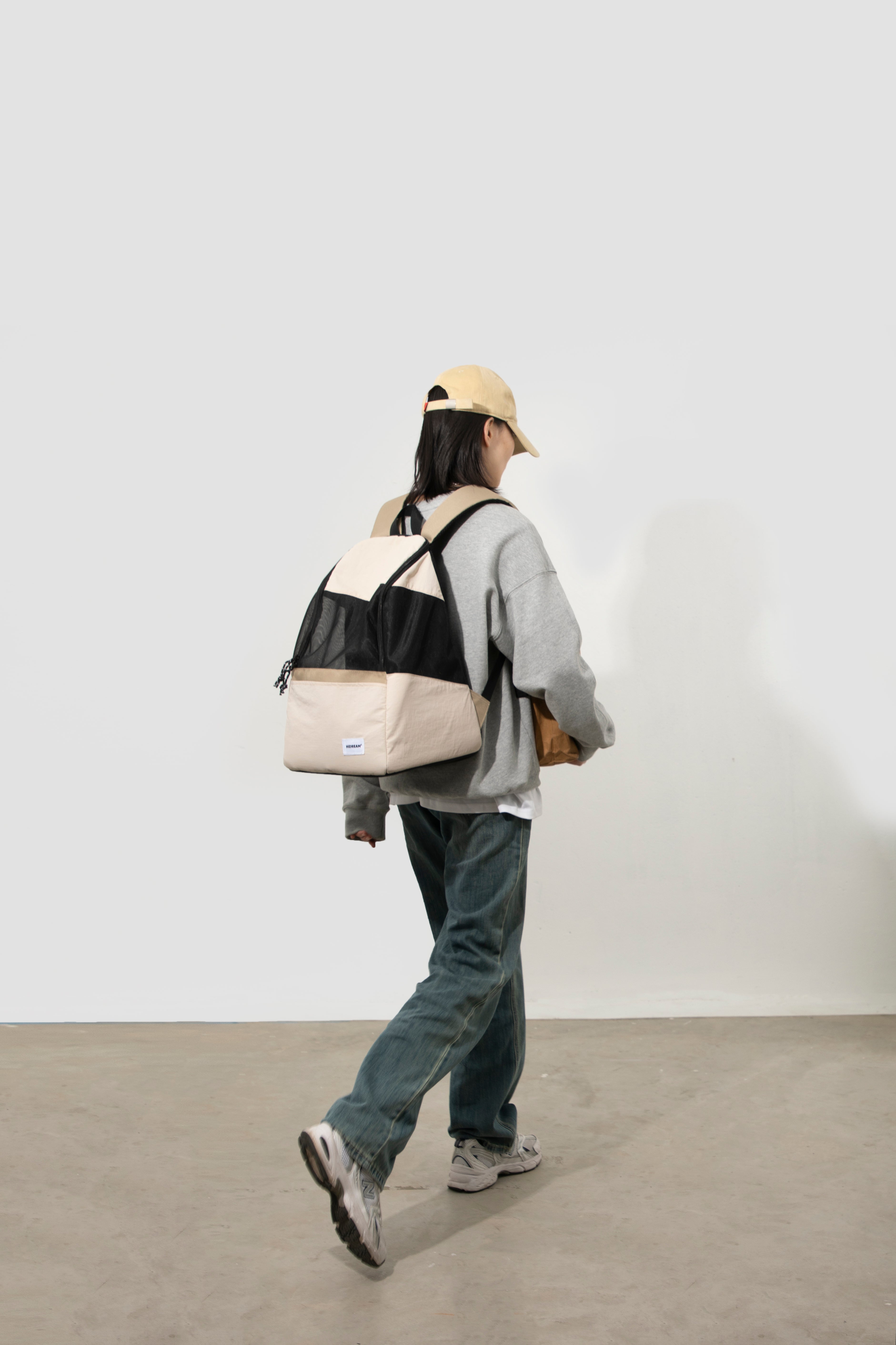 A person walking away in a minimalist studio setting, wearing a light beige baseball cap, a gray oversized sweatshirt, dark gray jeans, and sneakers, while carrying a color-blocked backpack with beige, black, and gray panels.