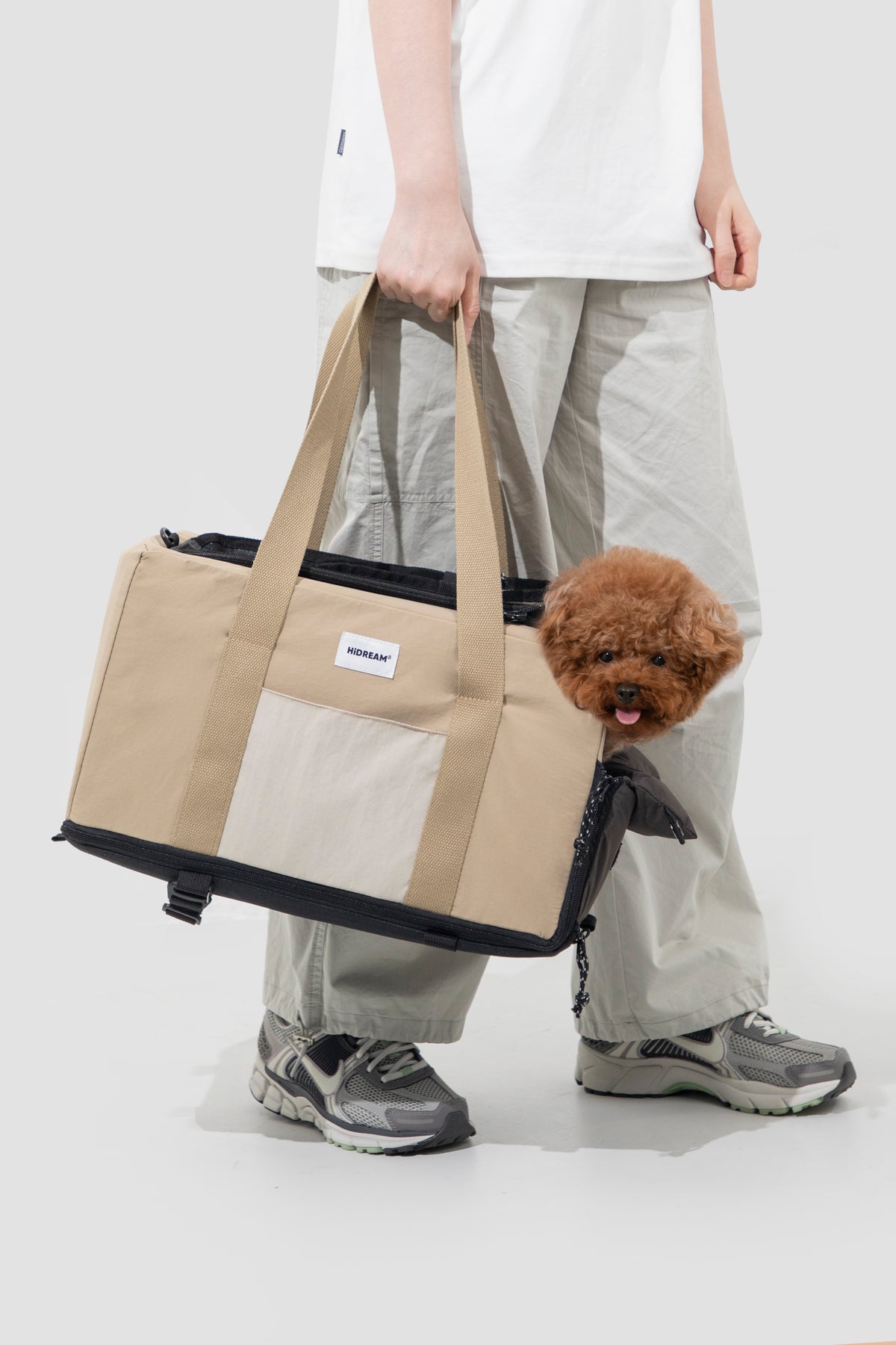 A beige and black pet carrier bag being carried by a person wearing a white t-shirt, light gray cargo pants, and gray sneakers, with a small brown poodle peeking out from the side of the bag.