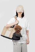 A person wearing a beige baseball cap, white t-shirt, and light gray pants carries a beige and black pet carrier bag with a small brown dog inside, against a light gray studio background.
