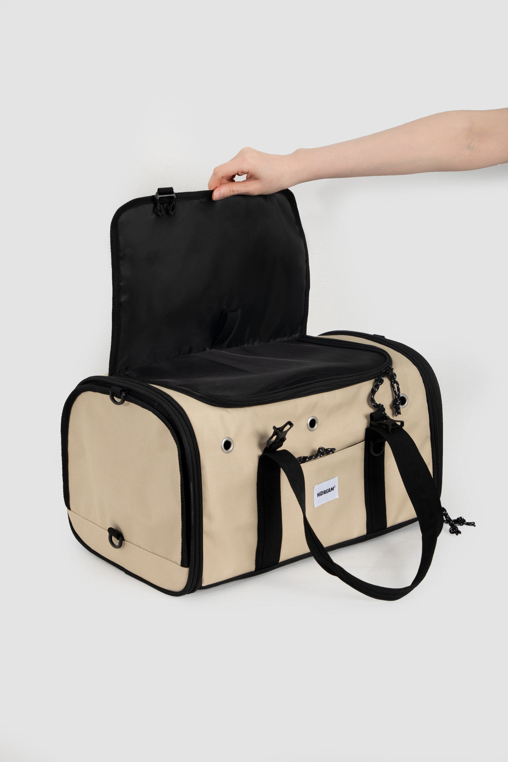 A beige and black pet carrier duffel bag with an open top, shown with a human arm reaching in to demonstrate the interior space, on a light gray background.