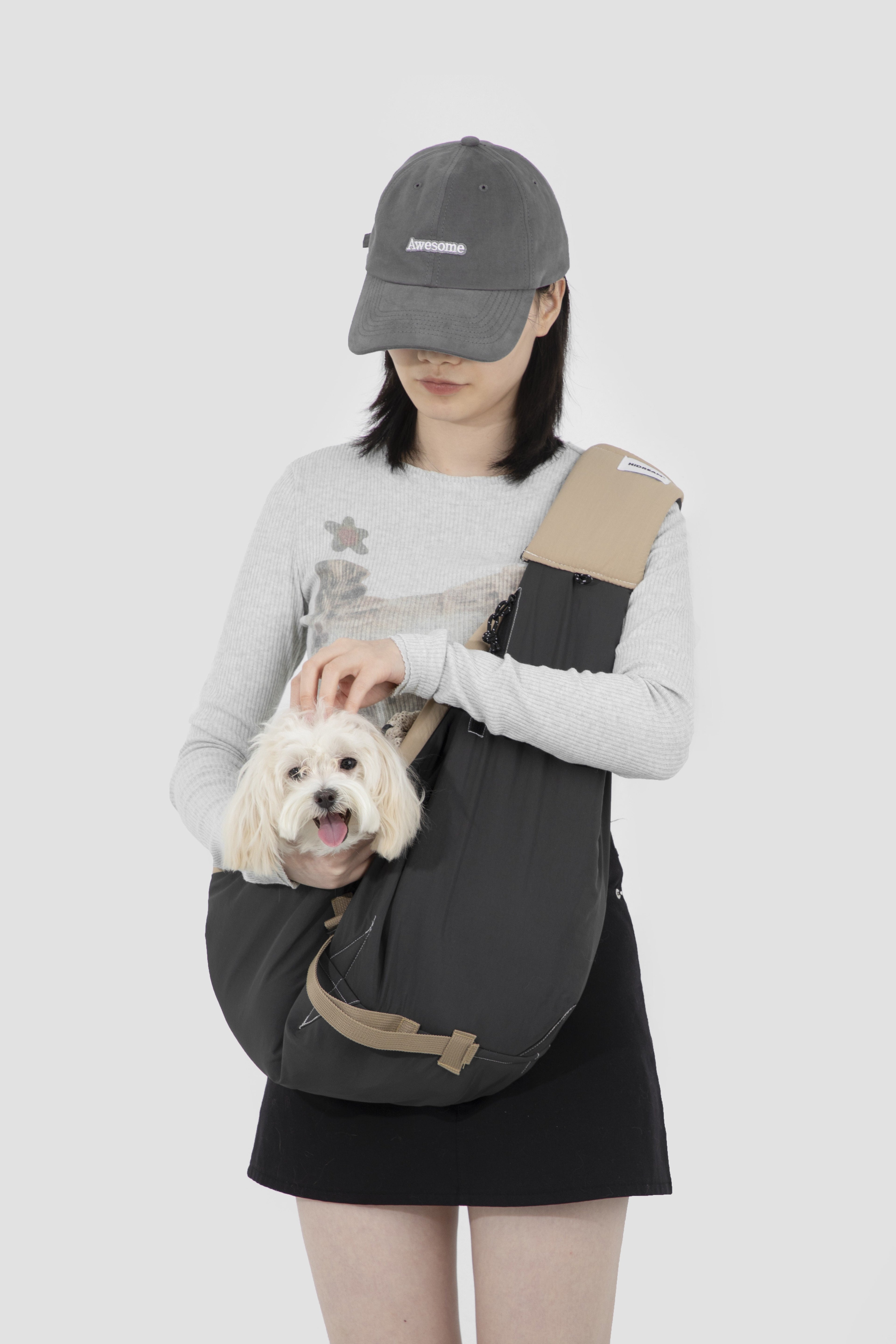 A woman wearing a gray baseball cap, light gray long-sleeve shirt, and black skirt models a dark gray pet carrier backpack with beige accents, holding a small white dog. The background is a solid light gray studio backdrop.