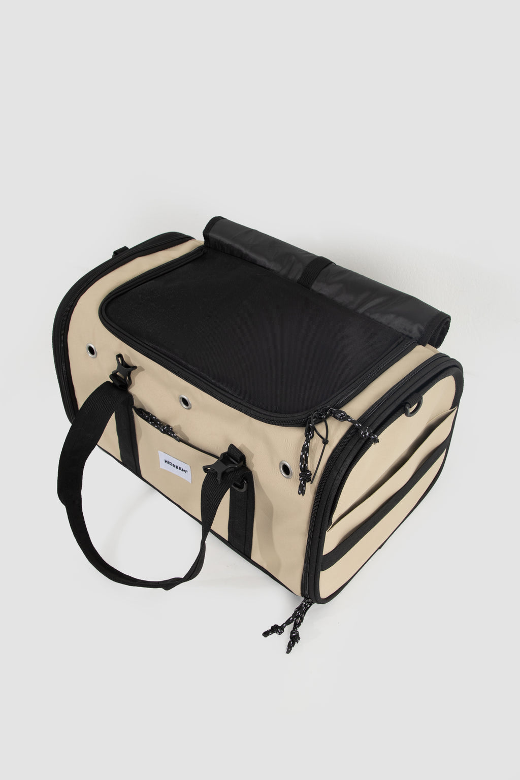 A beige and black pet carrier bag with a zippered top, side pockets, and a shoulder strap, photographed on a light gray background.