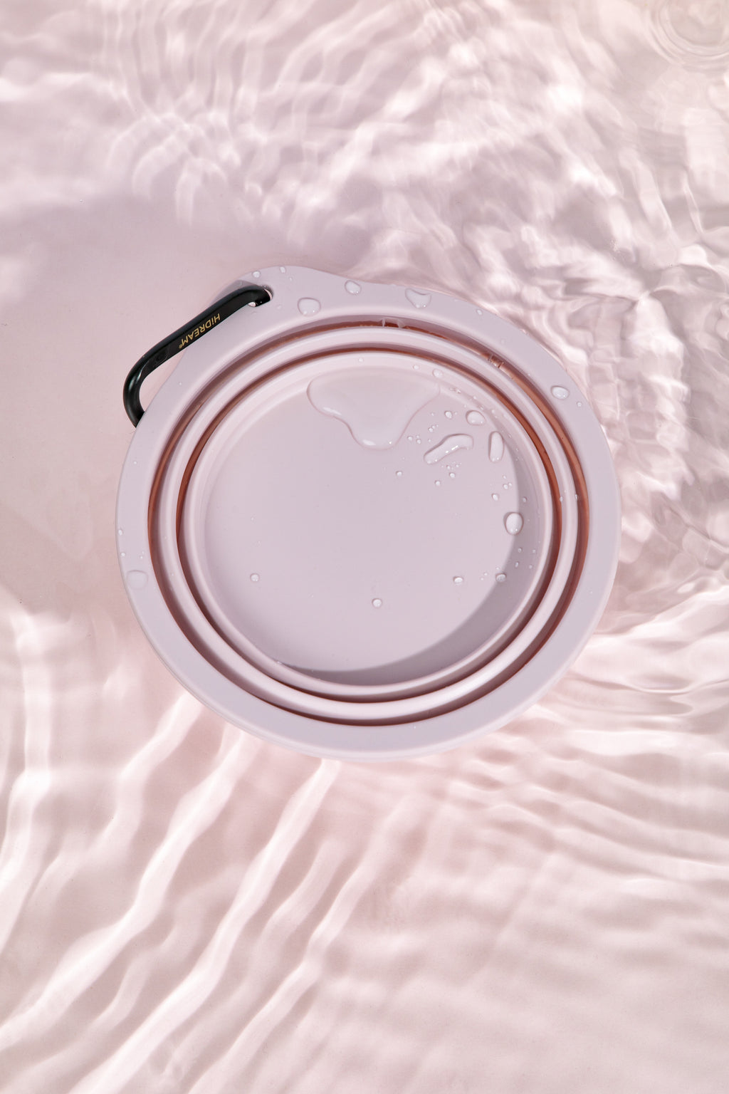 A top-down view of a white collapsible water bottle lid or cup insert with a black loop handle, shown partially filled with water and set against a soft pink water-textured background.