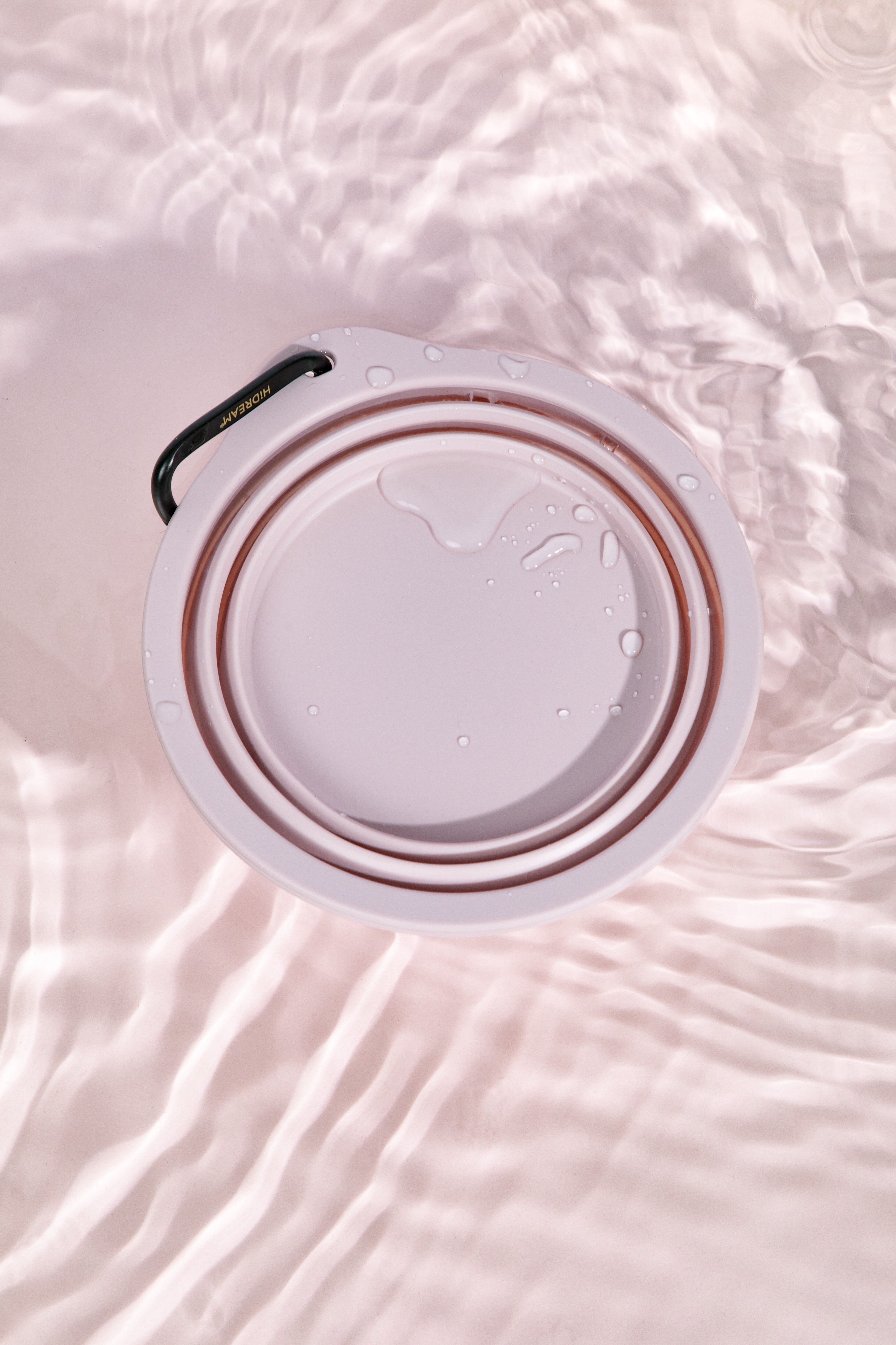 A top-down view of a white collapsible water bottle lid or cup insert with a black loop handle, shown partially filled with water and set against a soft pink water-textured background.