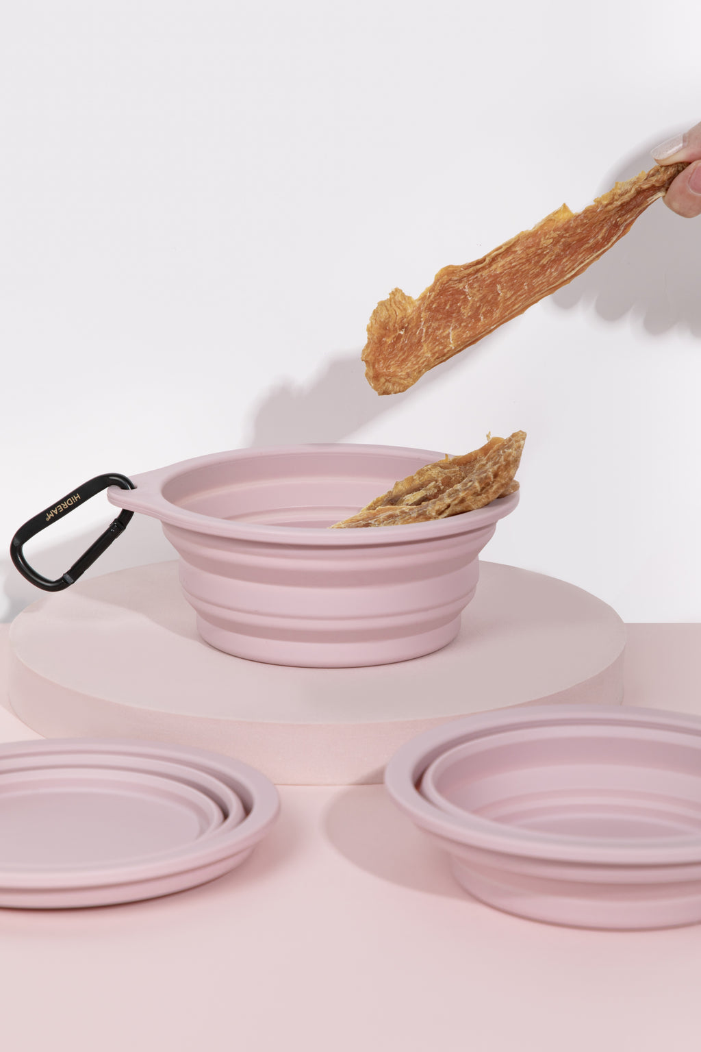 A pink collapsible silicone bowl with a black carabiner clip, shown with its matching lid and another folded bowl on a pink surface. A hand is holding a dried food strip above the bowl, and the background is a clean white studio setting.