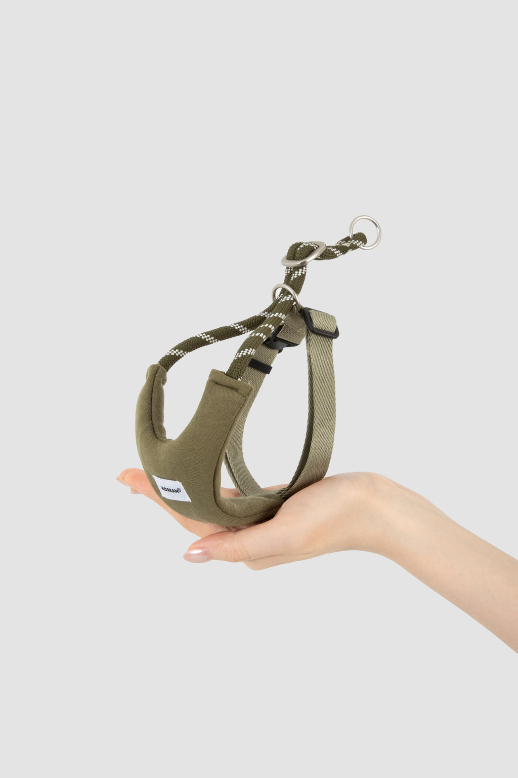 An olive green cat harness with a matching rope leash and metal ring, held in a person's hand against a light gray background.