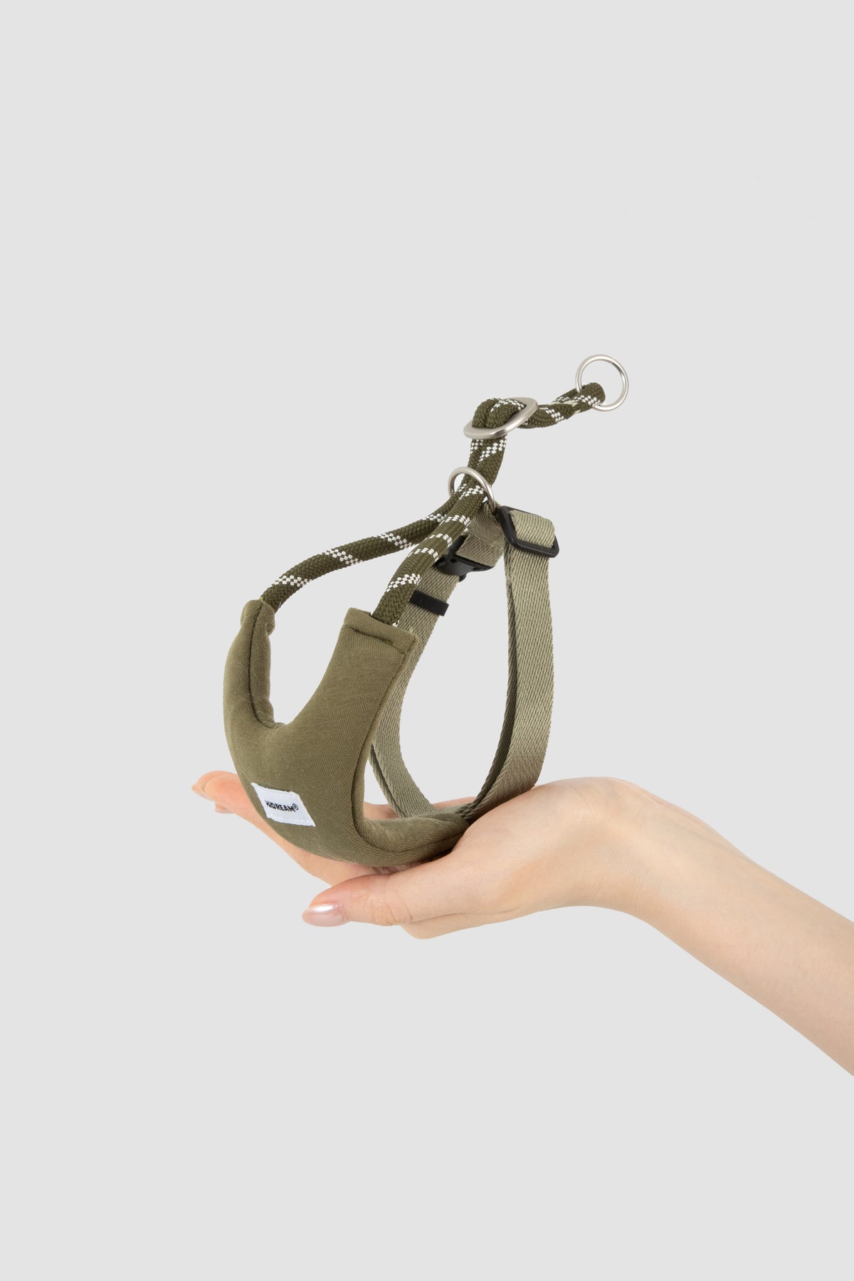 An olive green cat harness with a matching rope leash and metal ring, held in a person's hand against a light gray background.