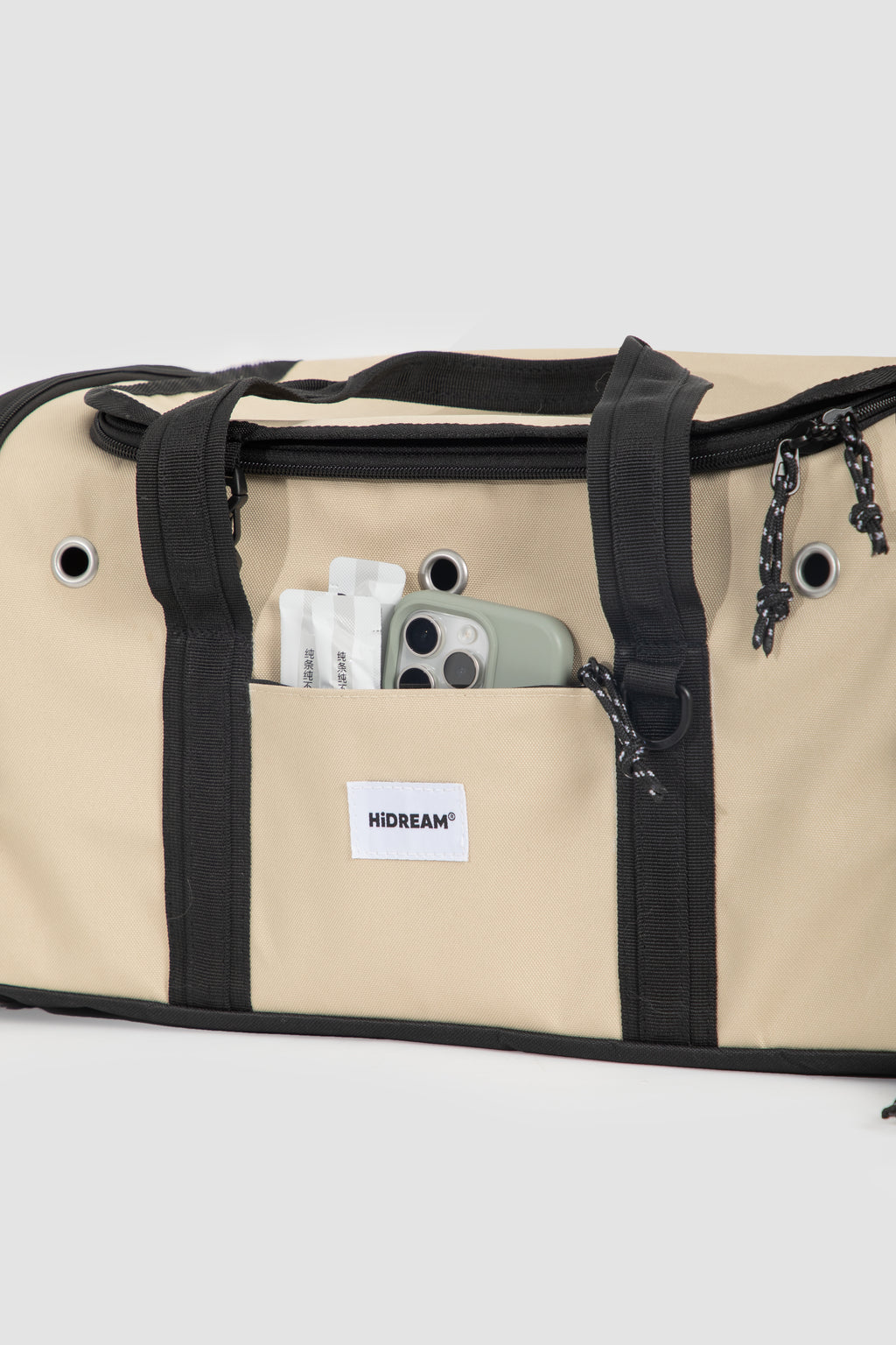 Beige canvas duffel bag with black trim, handles, and zipper details, featuring a front pocket with a visible HIDEAM brand label and a phone tucked inside.