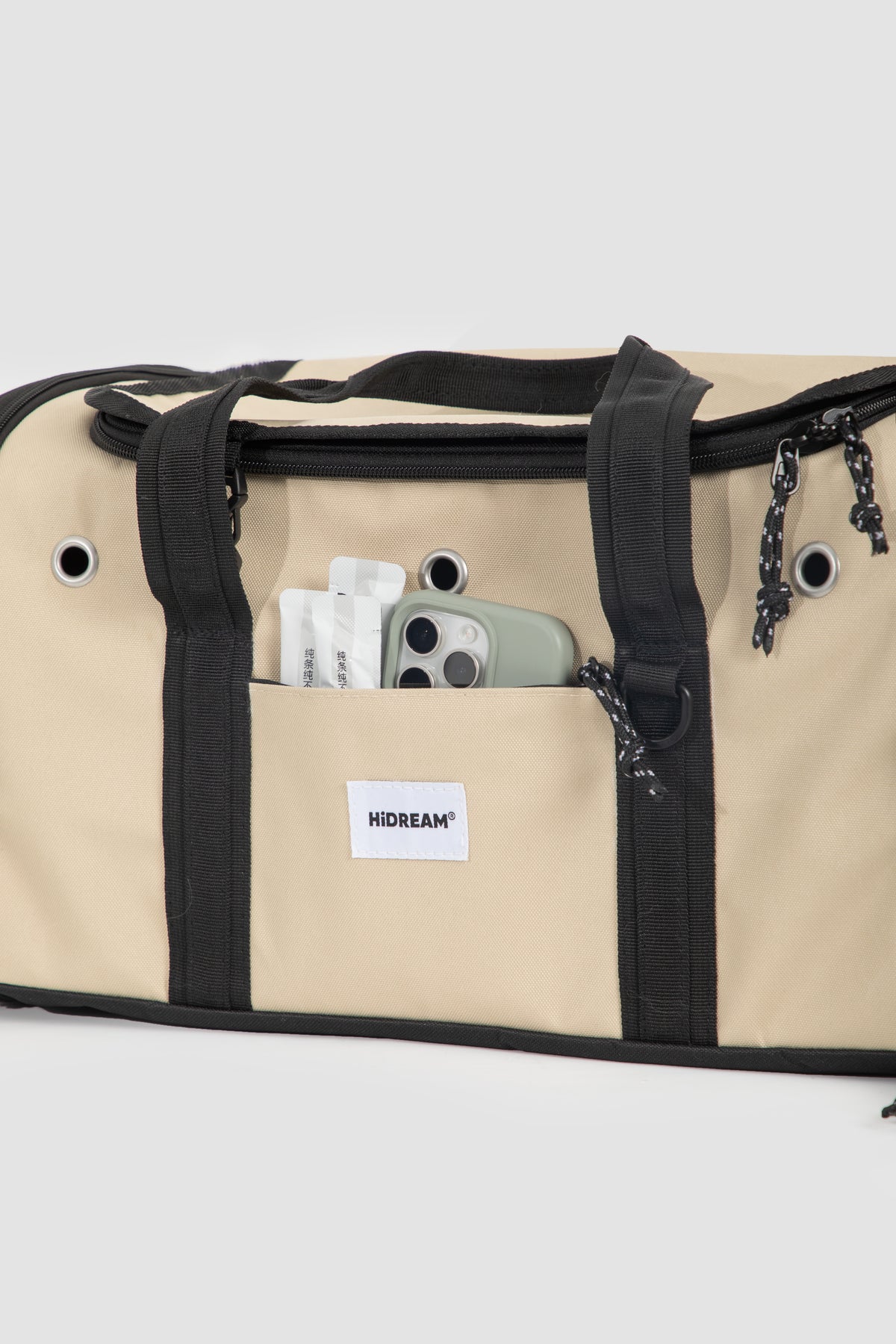 Beige canvas duffel bag with black trim, handles, and zipper details, featuring a front pocket with a visible HIDEAM brand label and a phone tucked inside.