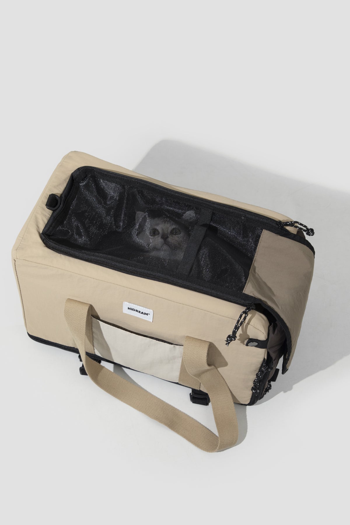 A beige and black pet carrier bag with a cat visible through its mesh window, photographed on a light gray studio background.