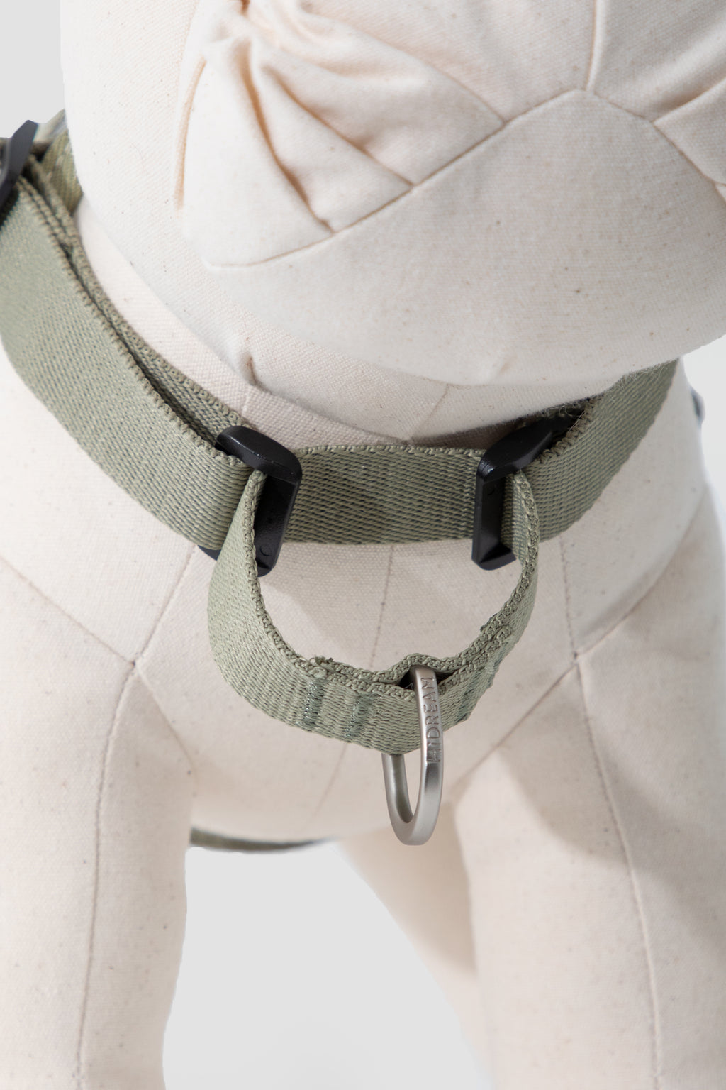 An olive green dog harness with black plastic adjusters and a silver metal ring, shown on a cream-colored dog mannequin.