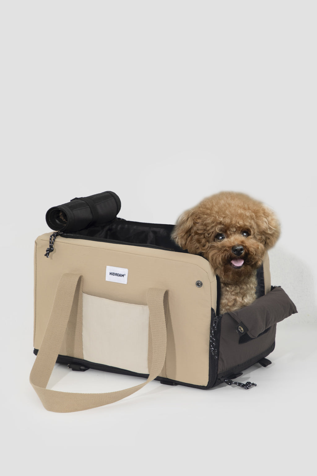 A small brown poodle sits in a beige and black pet carrier bag with a shoulder strap, photographed on a clean light gray studio background.