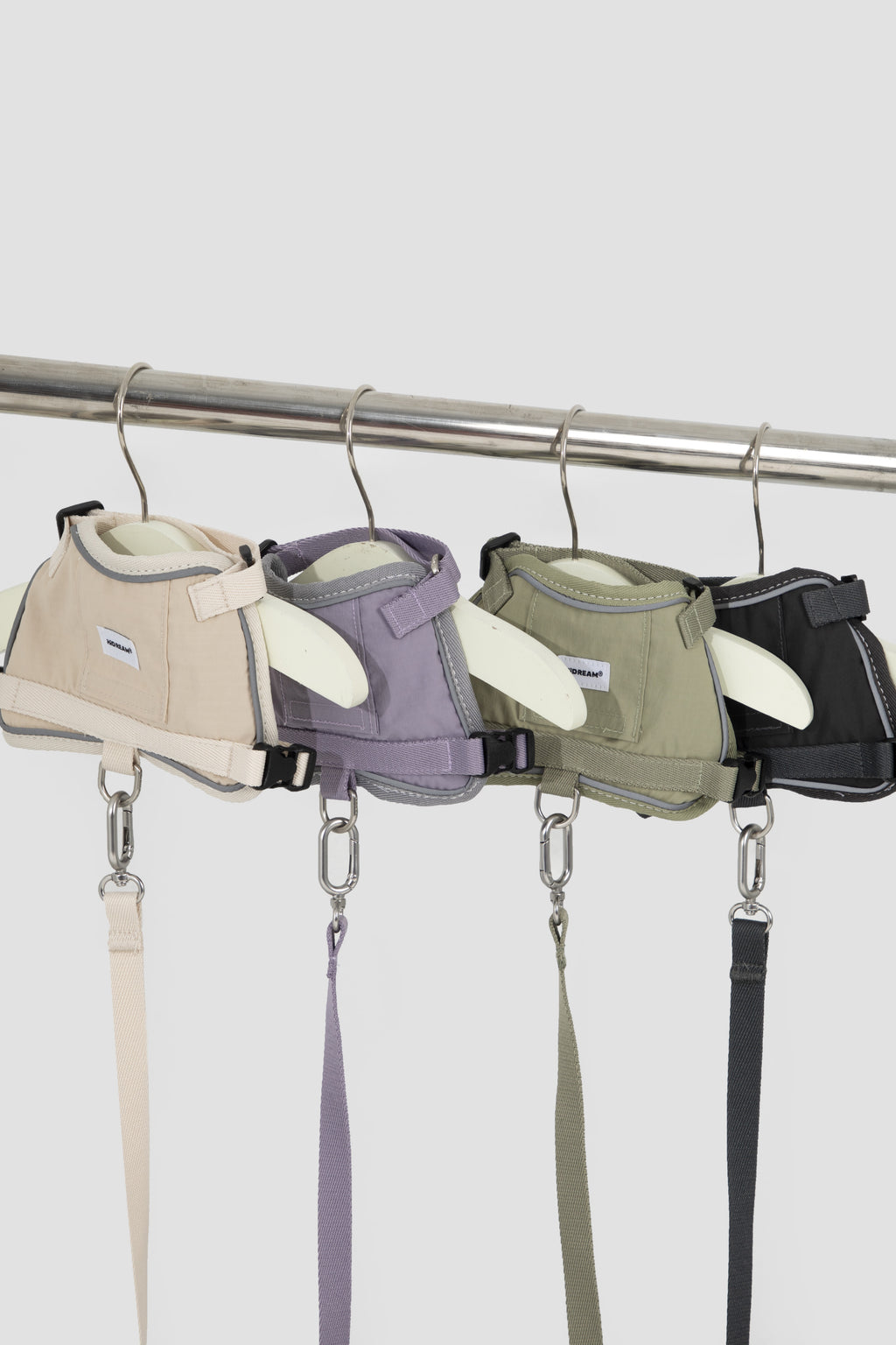 Four dog harnesses in beige, lavender, sage green, and black, hanging from a metal rack on a light gray background.