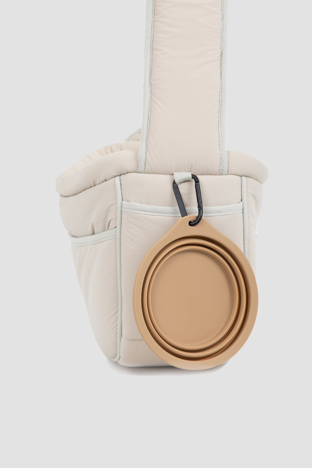 A cream-colored puffer-style shoulder bag with a tan collapsible silicone bowl clipped to the front, shown against a light gray studio background.