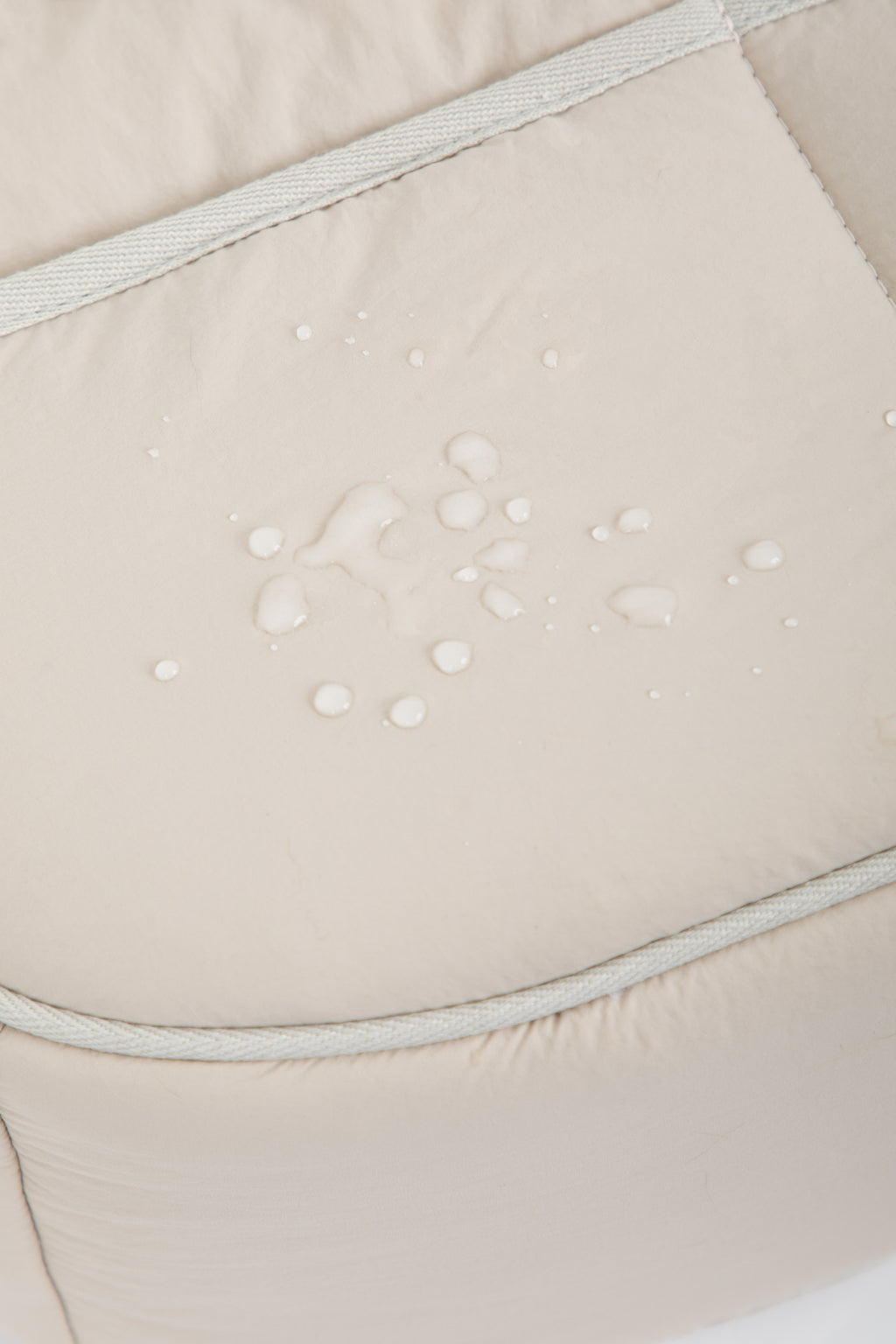 Close-up of a light beige padded surface with a few water droplets on it, showing a water-repellent finish. The fabric has subtle stitched paneling.