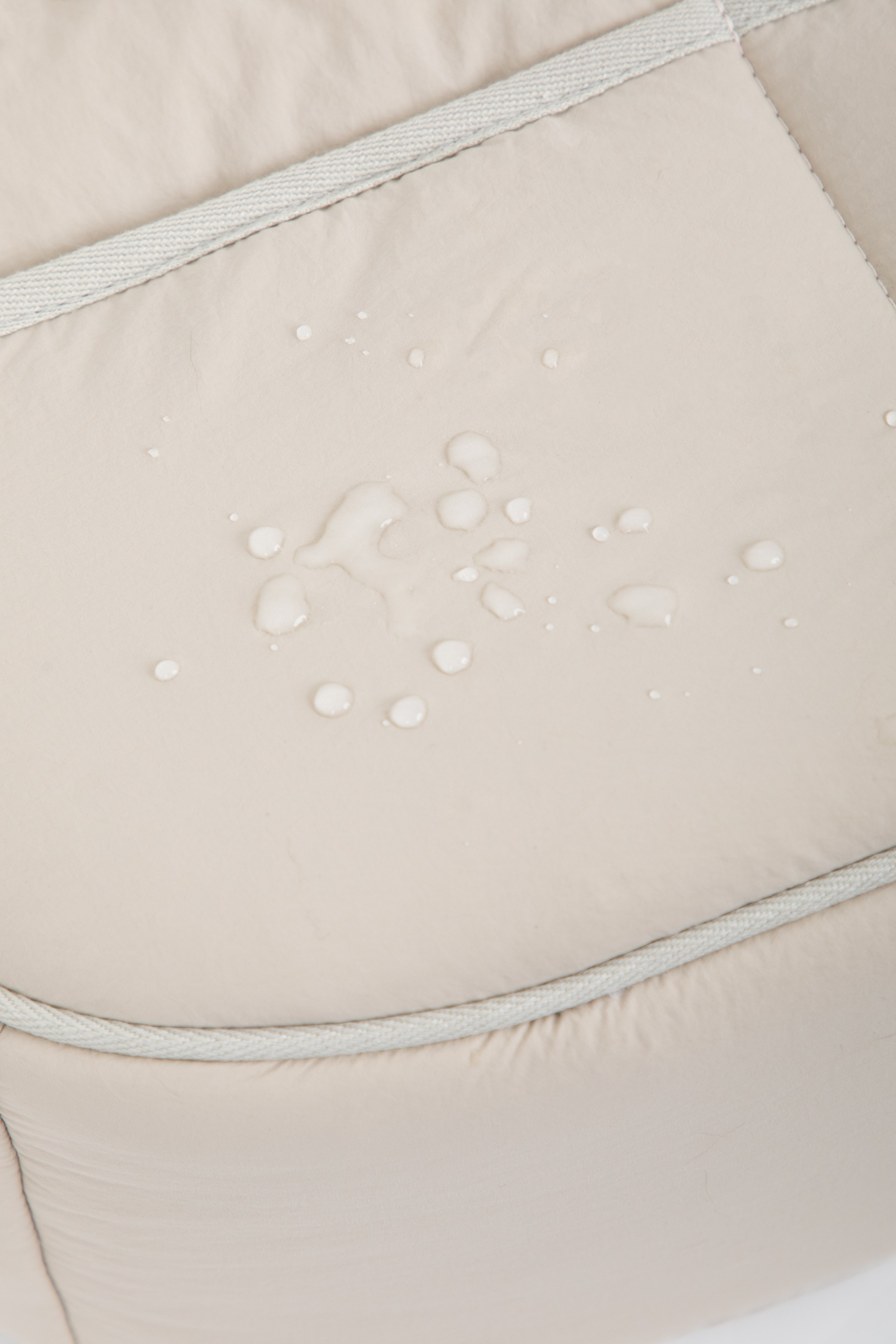 Close-up of a light beige padded surface with a few water droplets on it, showing a water-repellent finish. The fabric has subtle stitched paneling.
