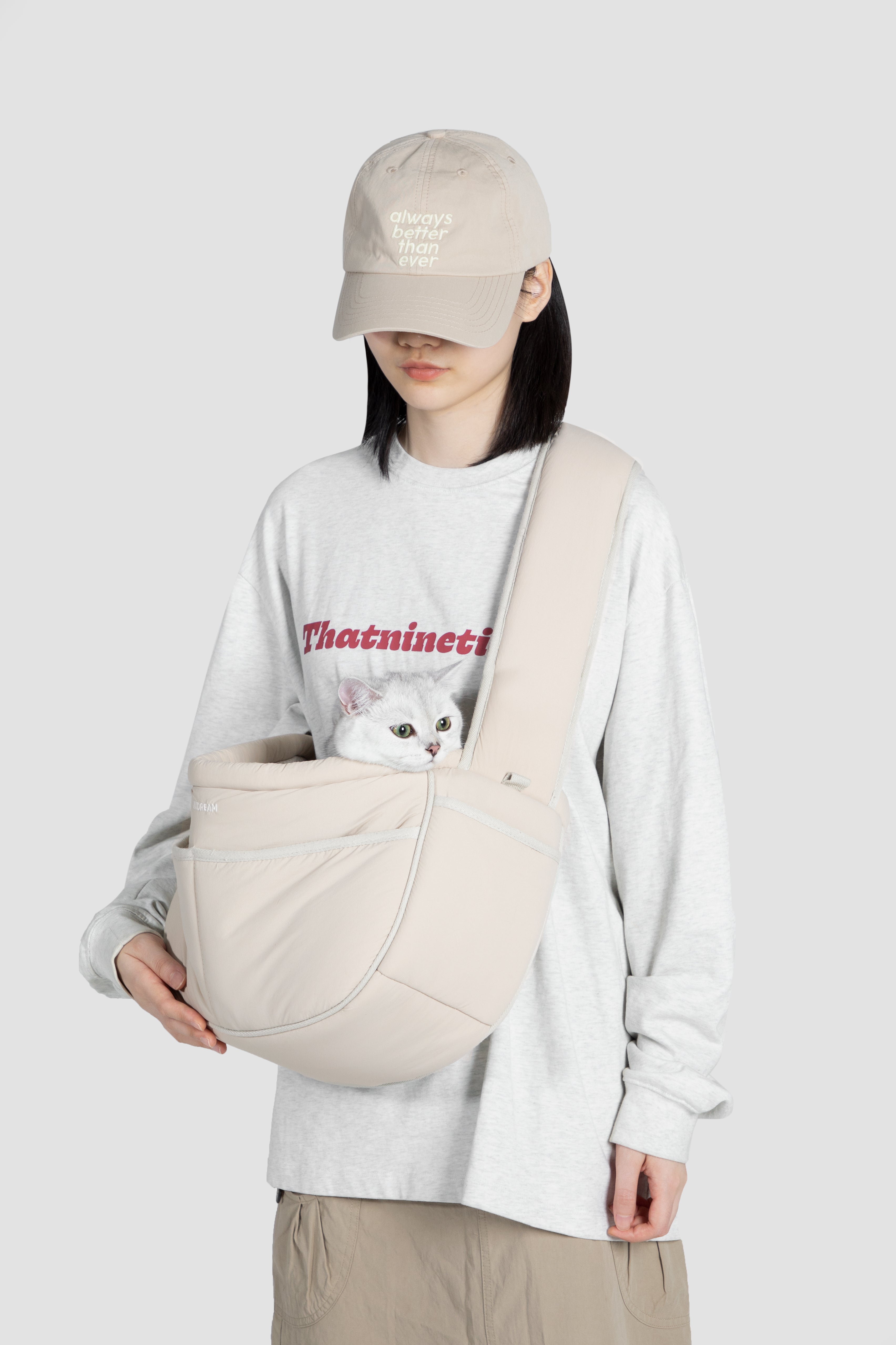 A model wearing a light gray sweatshirt, beige pants, and a beige baseball cap carries a white cat in a beige pet carrier bag worn across their chest. The background is a solid light gray studio backdrop.
