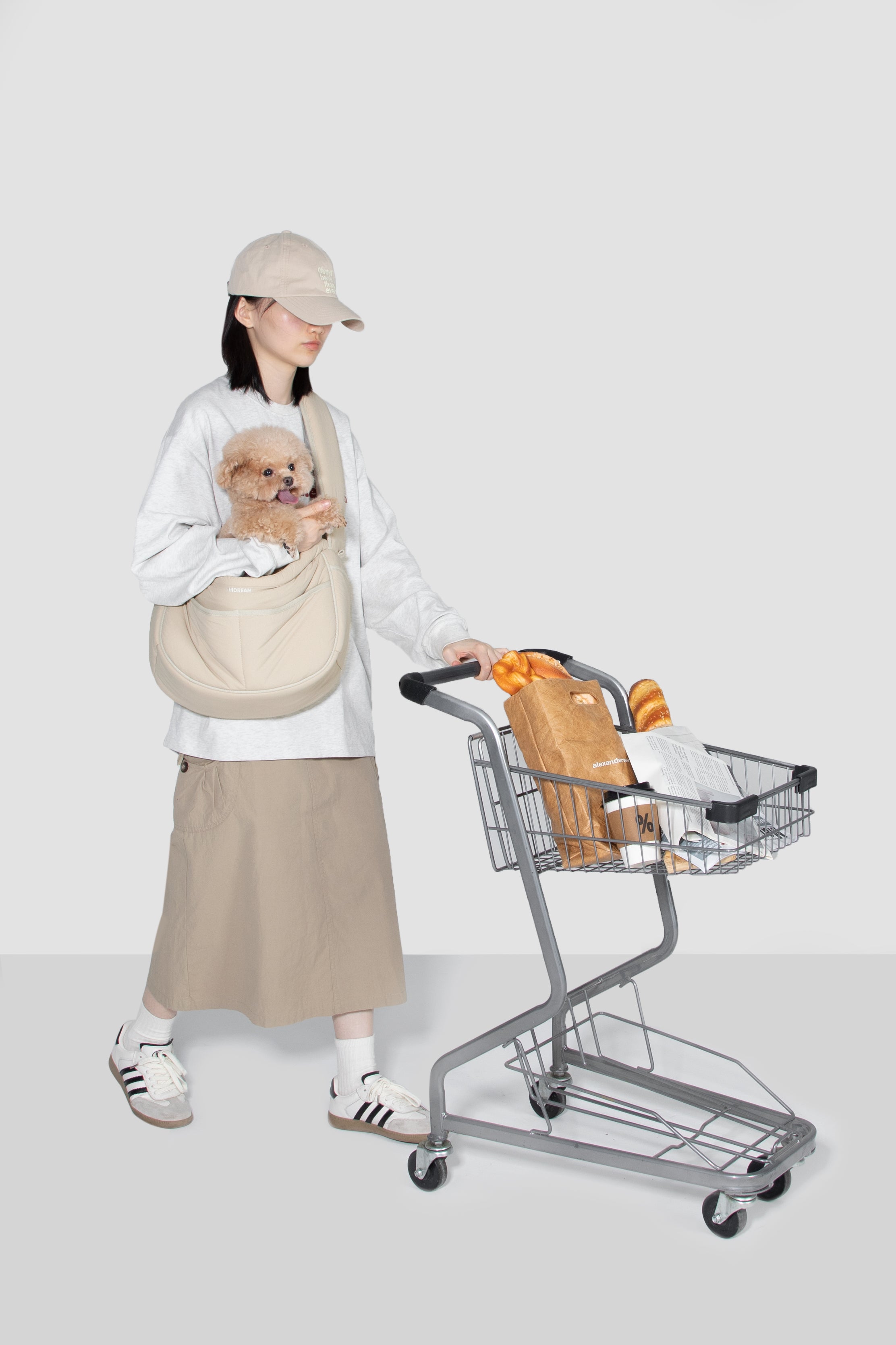 A woman wearing a beige cap, white sweatshirt, and beige skirt walks while pushing a metal shopping cart with a loaf of bread inside. She has a small light brown dog in a beige pet carrier bag worn crossbody.