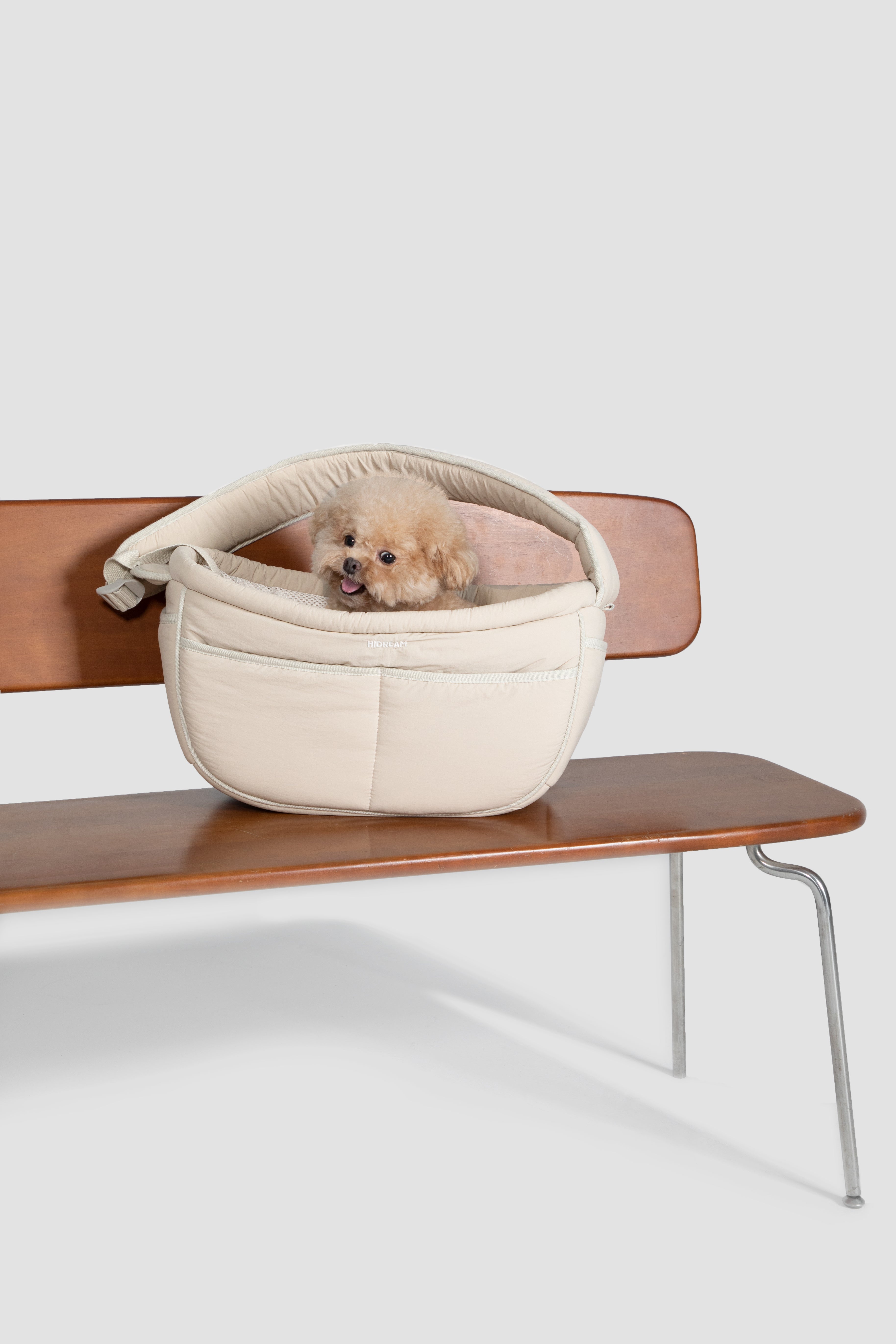 A small fluffy cream-colored dog sits inside a cream-colored pet bed placed on a mid-century modern wooden bench against a light gray background.
