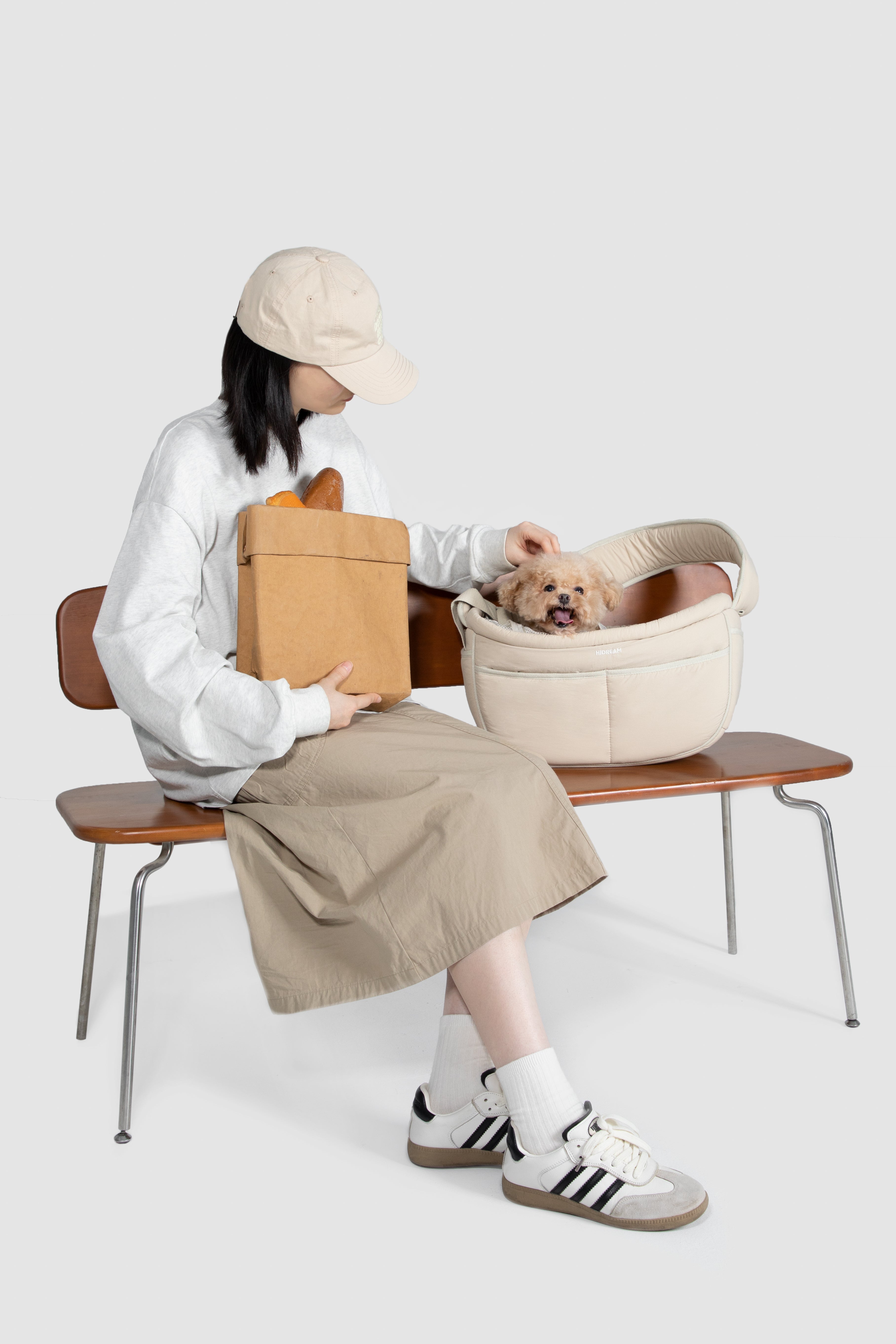A woman sits on a wooden bench holding a brown paper bag next to a cream-colored pet basket containing a small dog, against a solid light gray background.