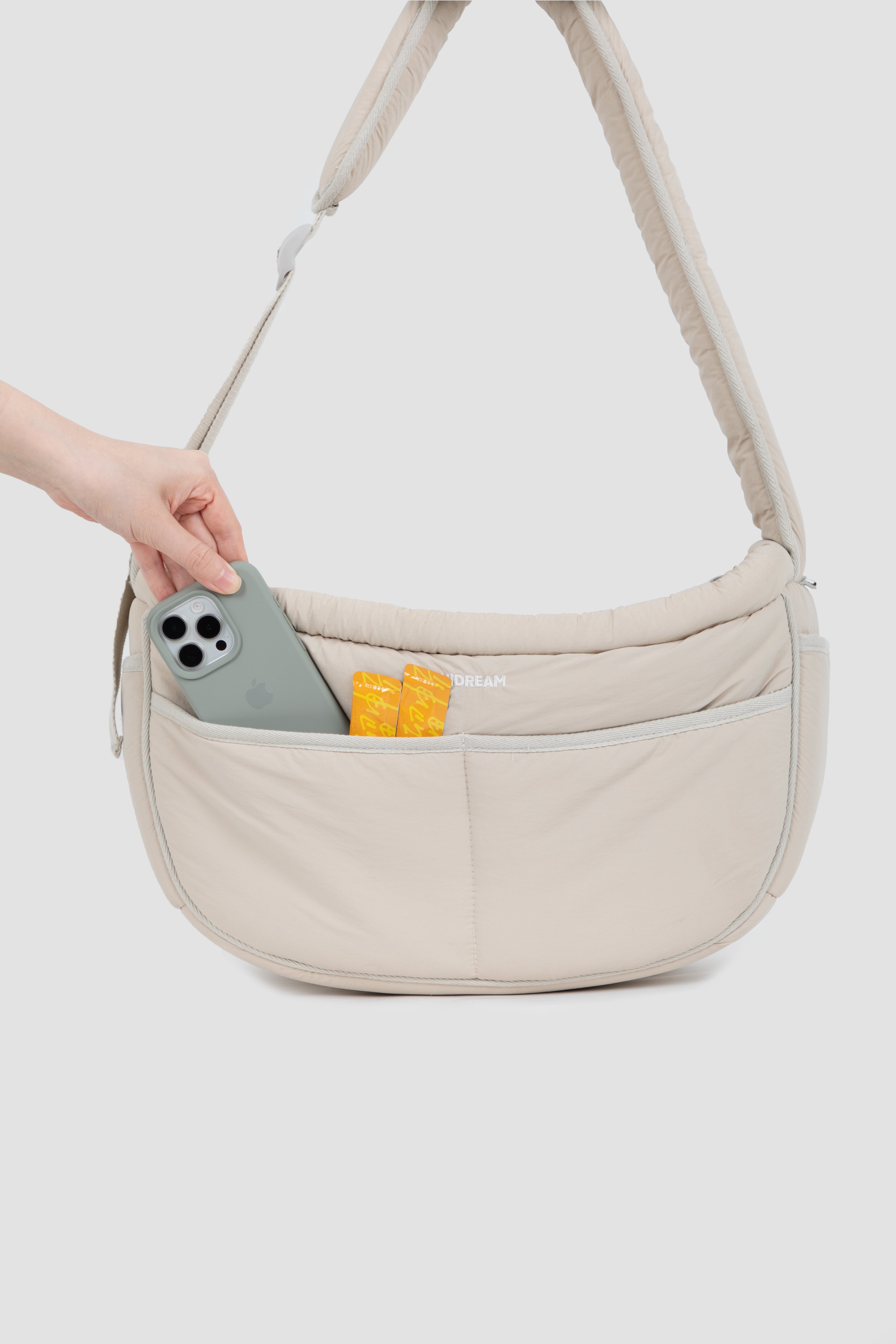 Beige crossbody bag with a pocket holding a phone and snacks on a light gray background