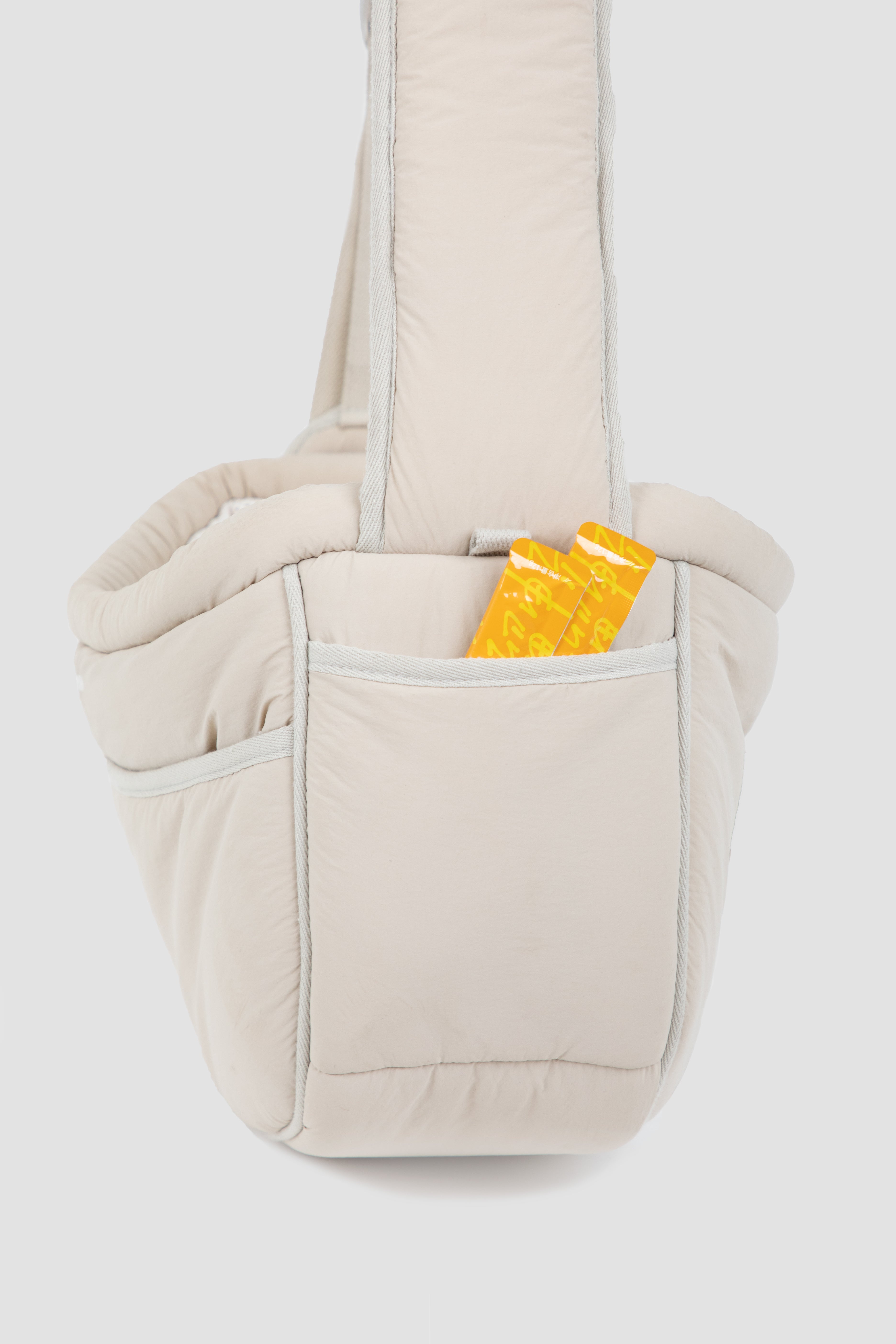 A light beige quilted baby carrier bag with a wide shoulder strap, featuring a front pocket holding a yellow patterned item, shown against a light gray background.