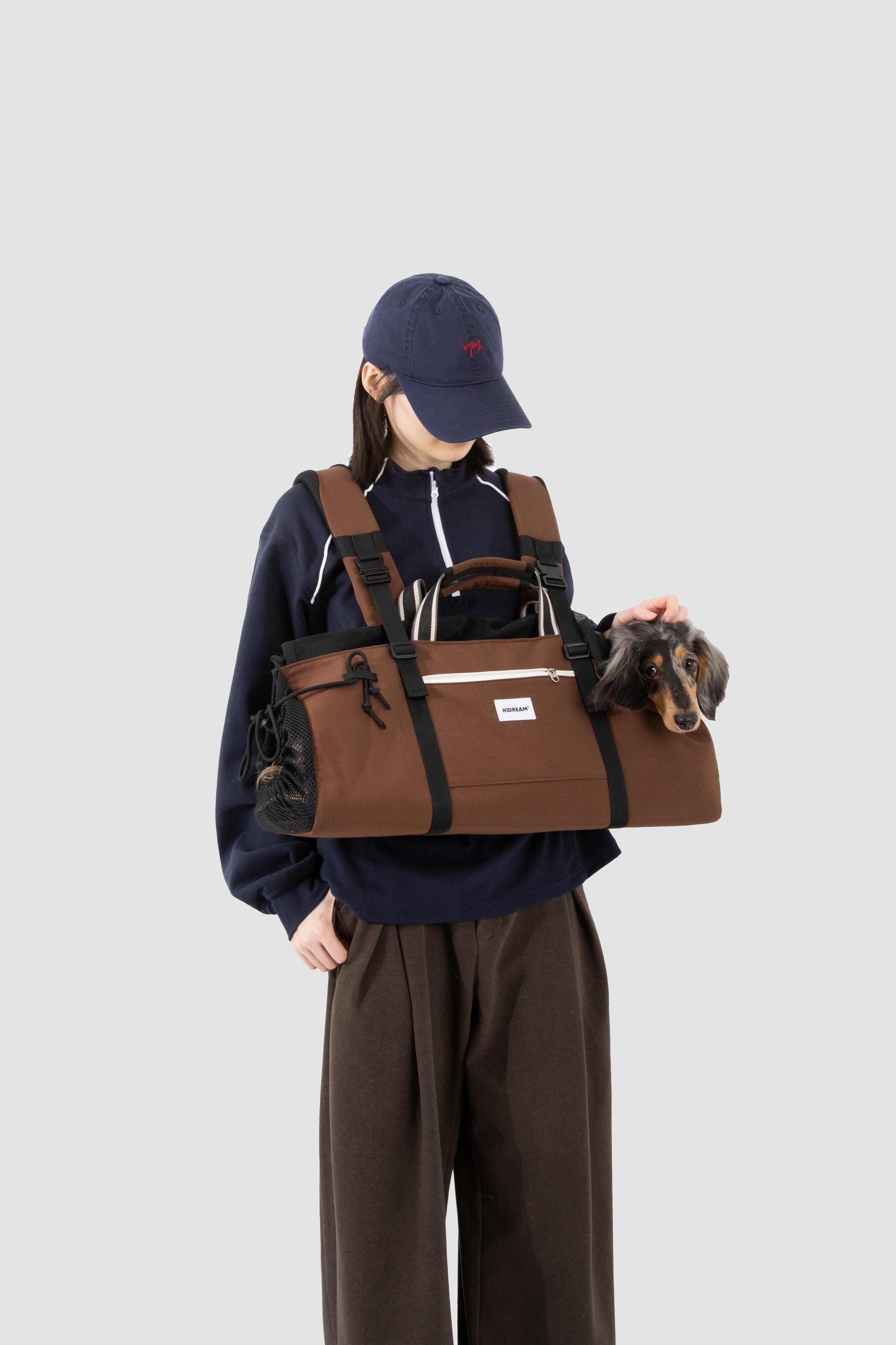 A person wearing a navy blue cap, navy zip-up hoodie, and dark brown wide-leg pants carries a brown pet carrier duffel bag with a Dachshund inside, against a solid light gray background.