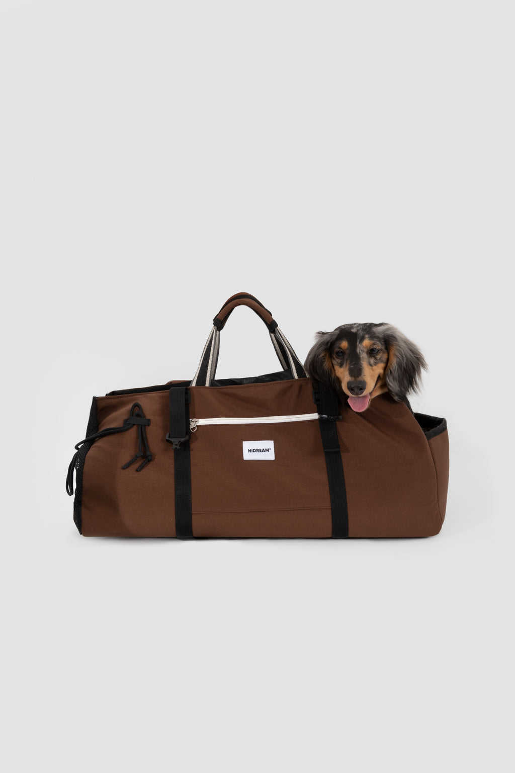 A brown duffel bag with black straps and handles, featuring a small white rectangular logo patch on the front. A dachshund dog is peeking out from the top right of the bag.