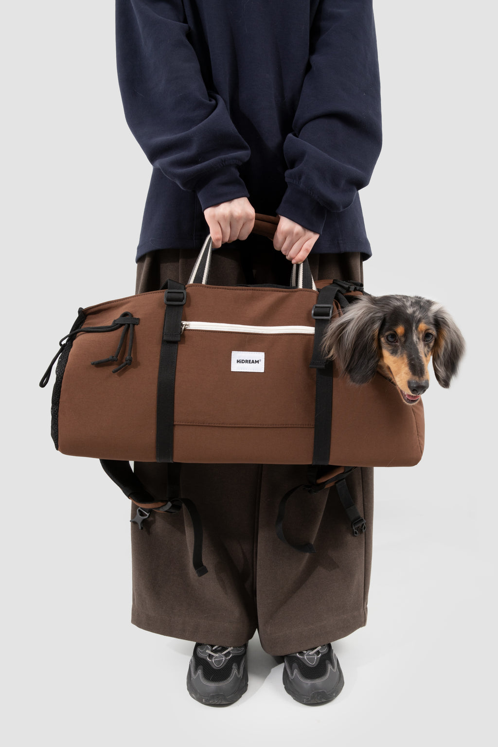 A person wearing a navy sweatshirt and brown pants holds a brown duffle bag with a small dachshund dog peeking out from the side. The bag has black straps, a white horizontal stripe, and a small white brand label on the front.