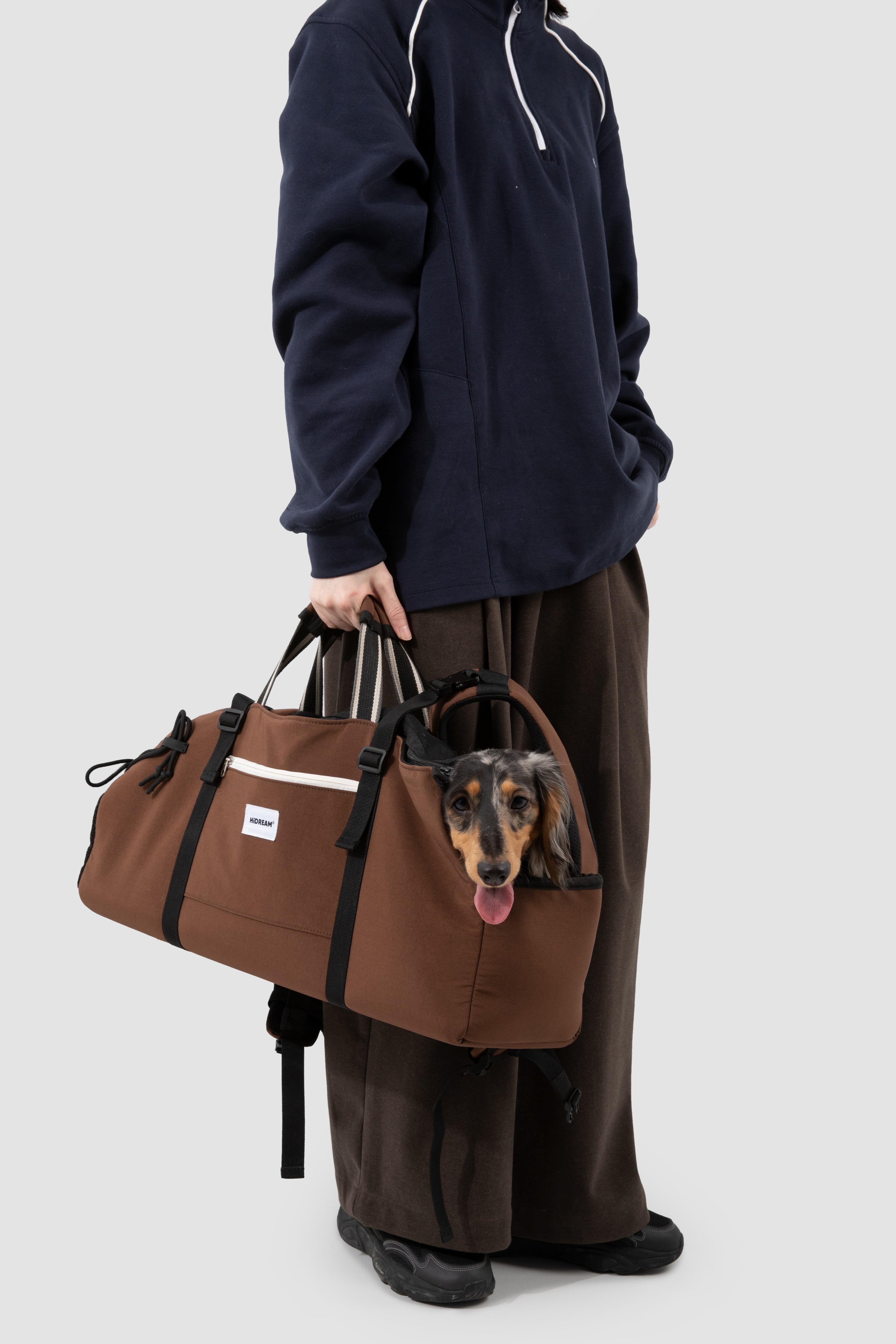 A person wearing a navy hoodie and brown pants carries a brown duffle bag with a dachshund dog peeking out from the top on a light gray background.