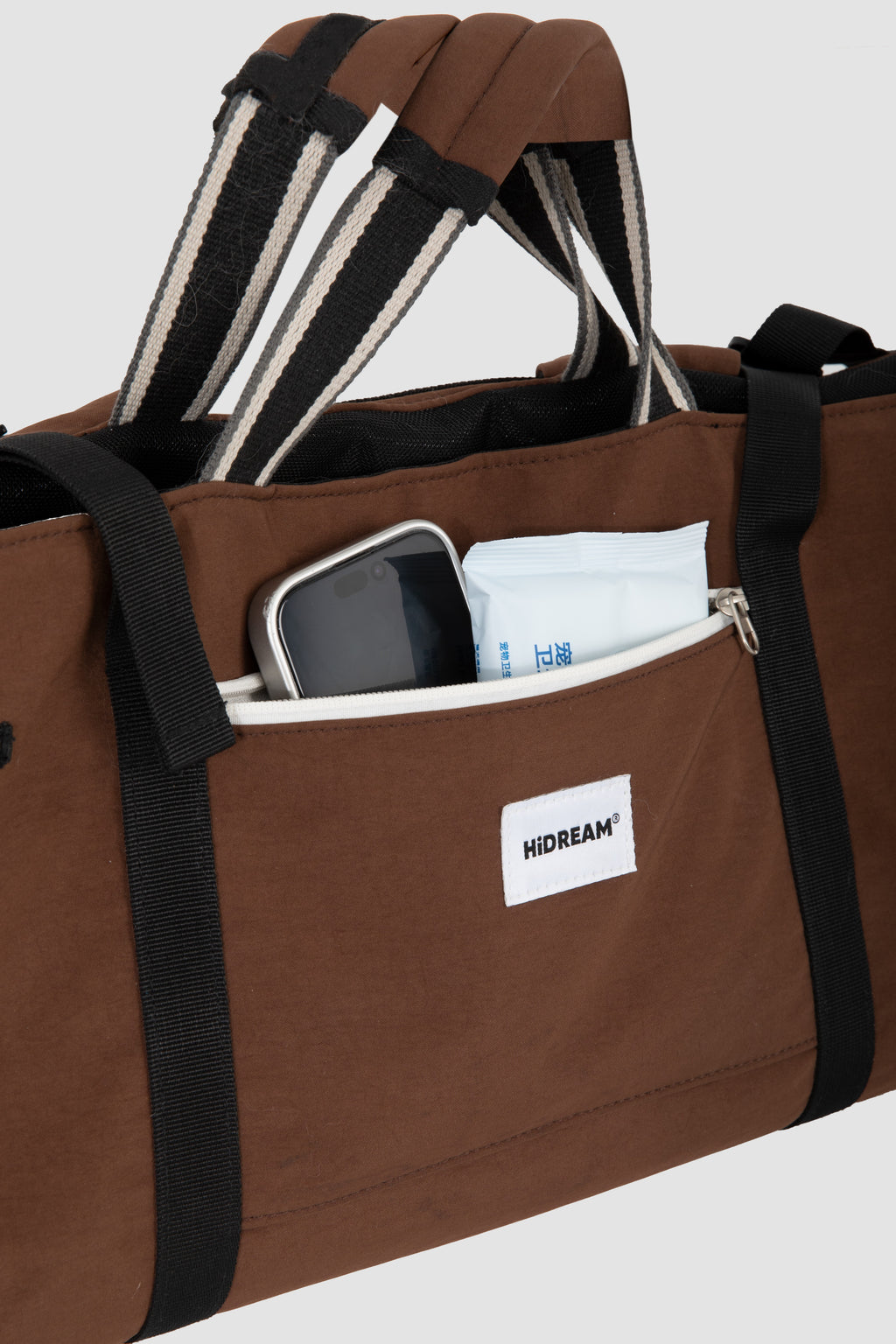 A brown Dachshund bag with black and white striped handles, shown with a smartphone and white packet in its front pocket. A white label on the front pocket displays the brand 'HIDREAM'.