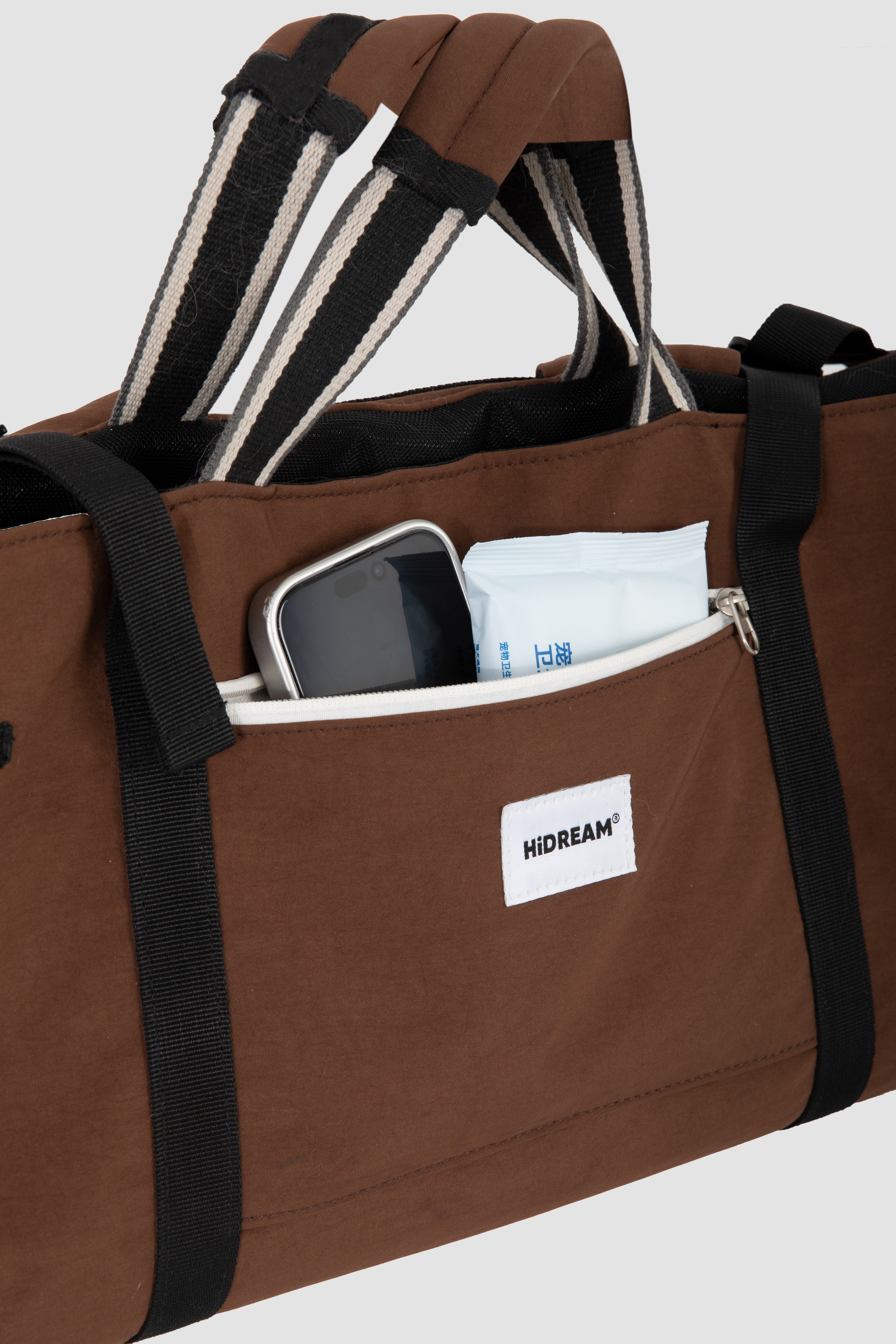 A brown Dachshund bag with black and white striped handles, shown with a smartphone and white packet in its front pocket. A white label on the front pocket displays the brand 'HIDREAM'.