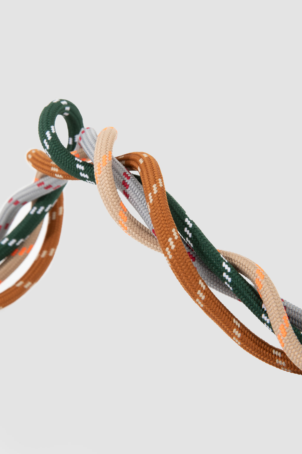 A close-up of twisted elastic cords or straps in multiple colors, including dark green, tan, beige, and gray, with small white dotted details, set against a solid light gray background.