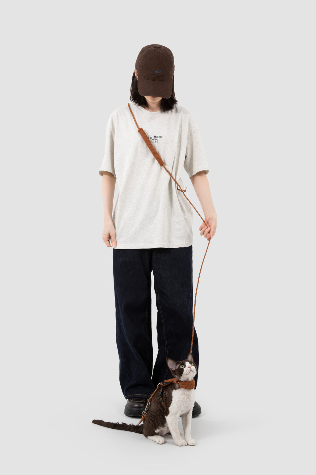 A person wearing a brown baseball cap, cream-colored t-shirt, and dark wide-leg pants stands holding a brown leather dog leash attached to a cat, against a solid light gray background.