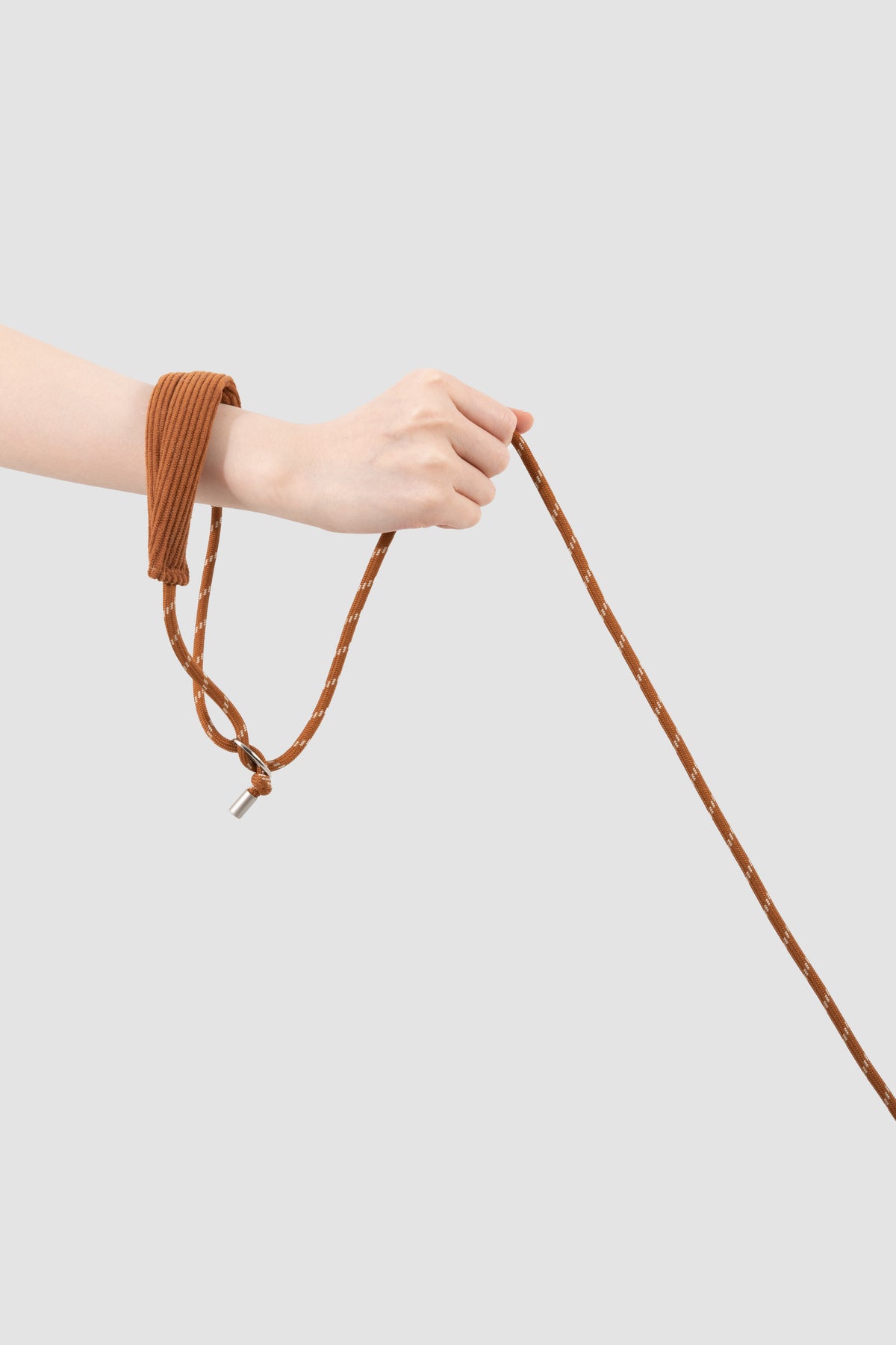 A person's hand and forearm wearing a brown ribbed fabric wrist strap attached to a matching brown rope, shown against a light gray studio background.